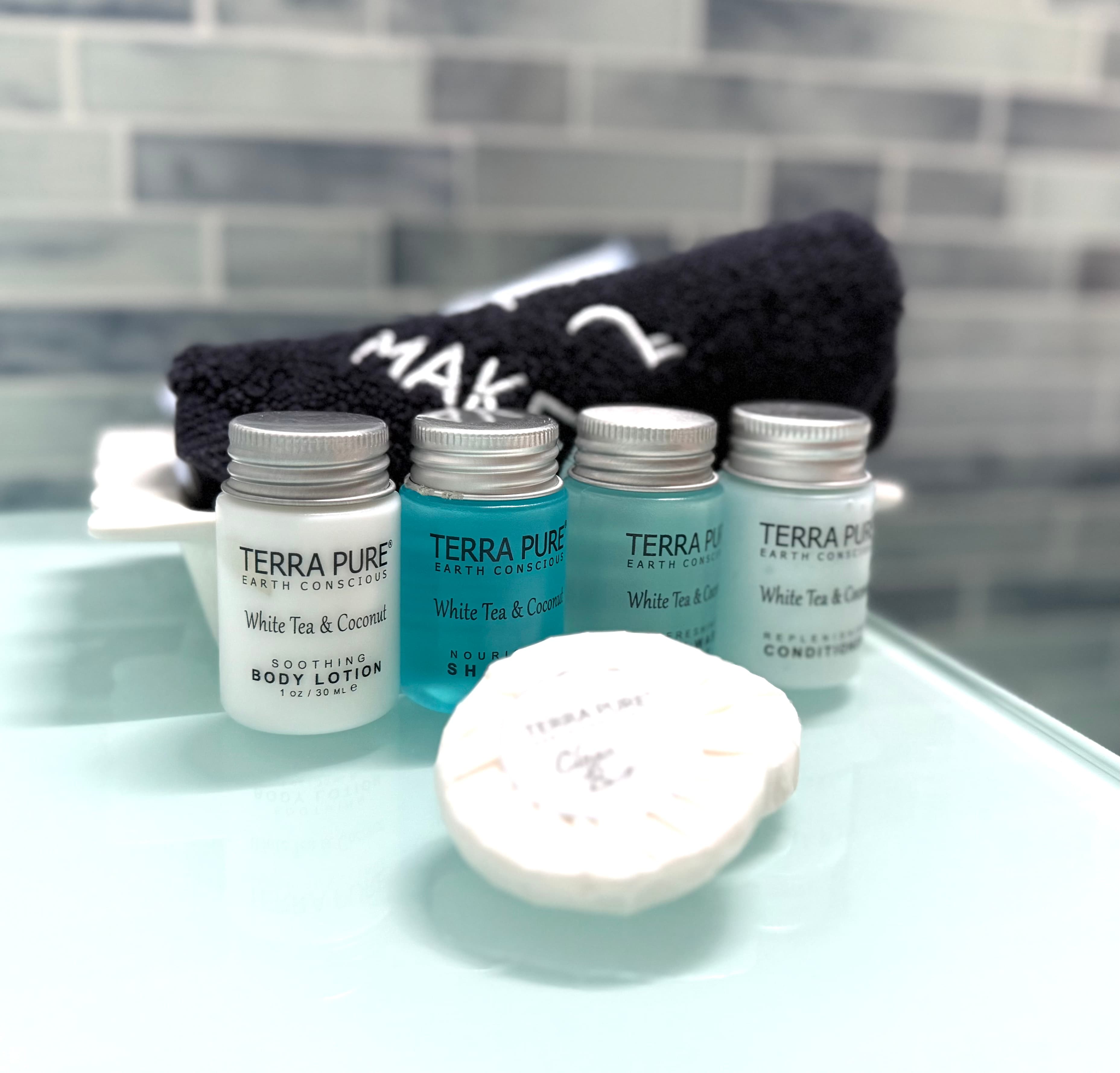 Four small bottles of Terra Pure white tea and coconut body lotion, shampoo, and conditioner, alongside a round soap and a black towel with "MAKE" written on it.