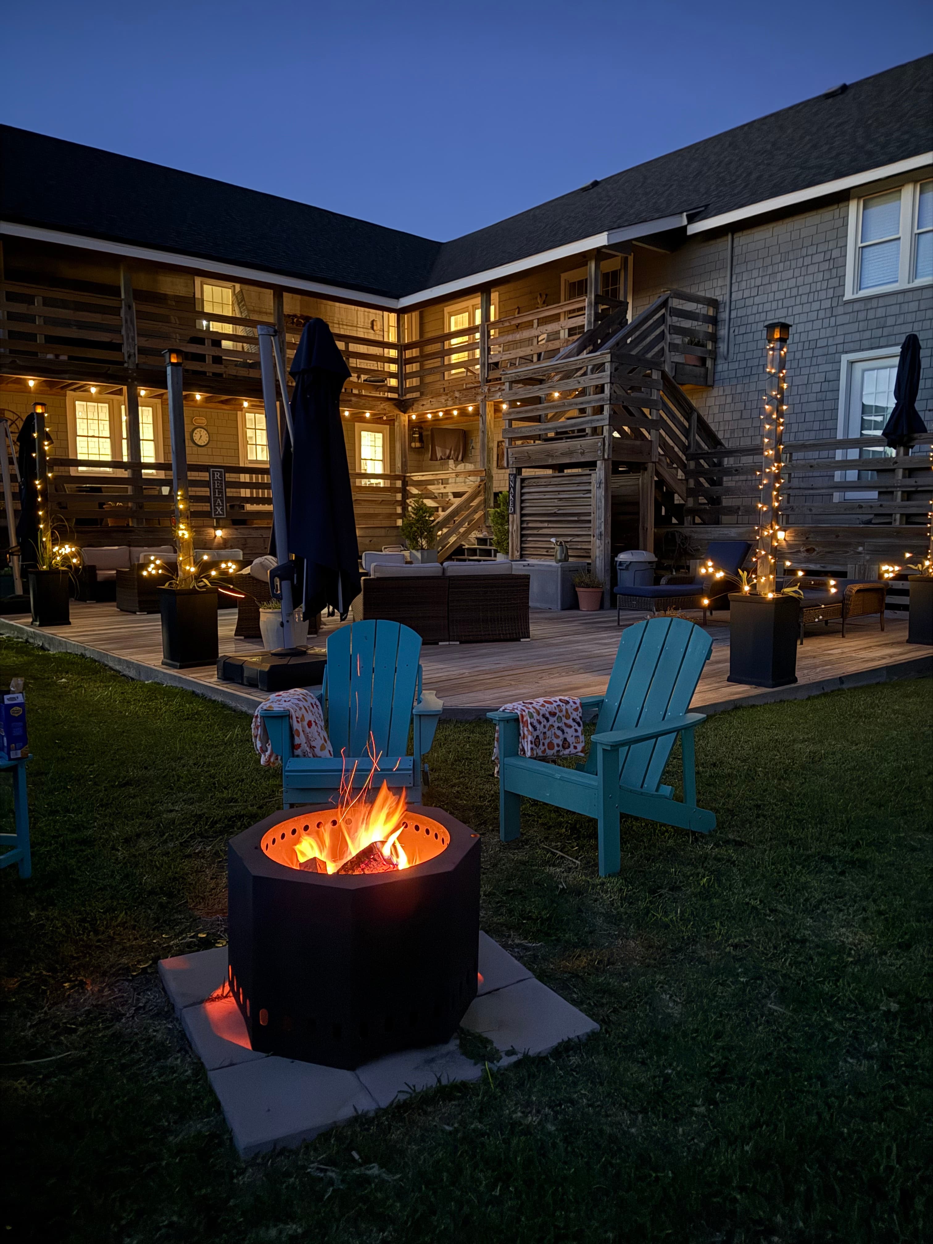 A cozy backyard fire pit surrounded by teal chairs, with warm lights adorning the patio and building.