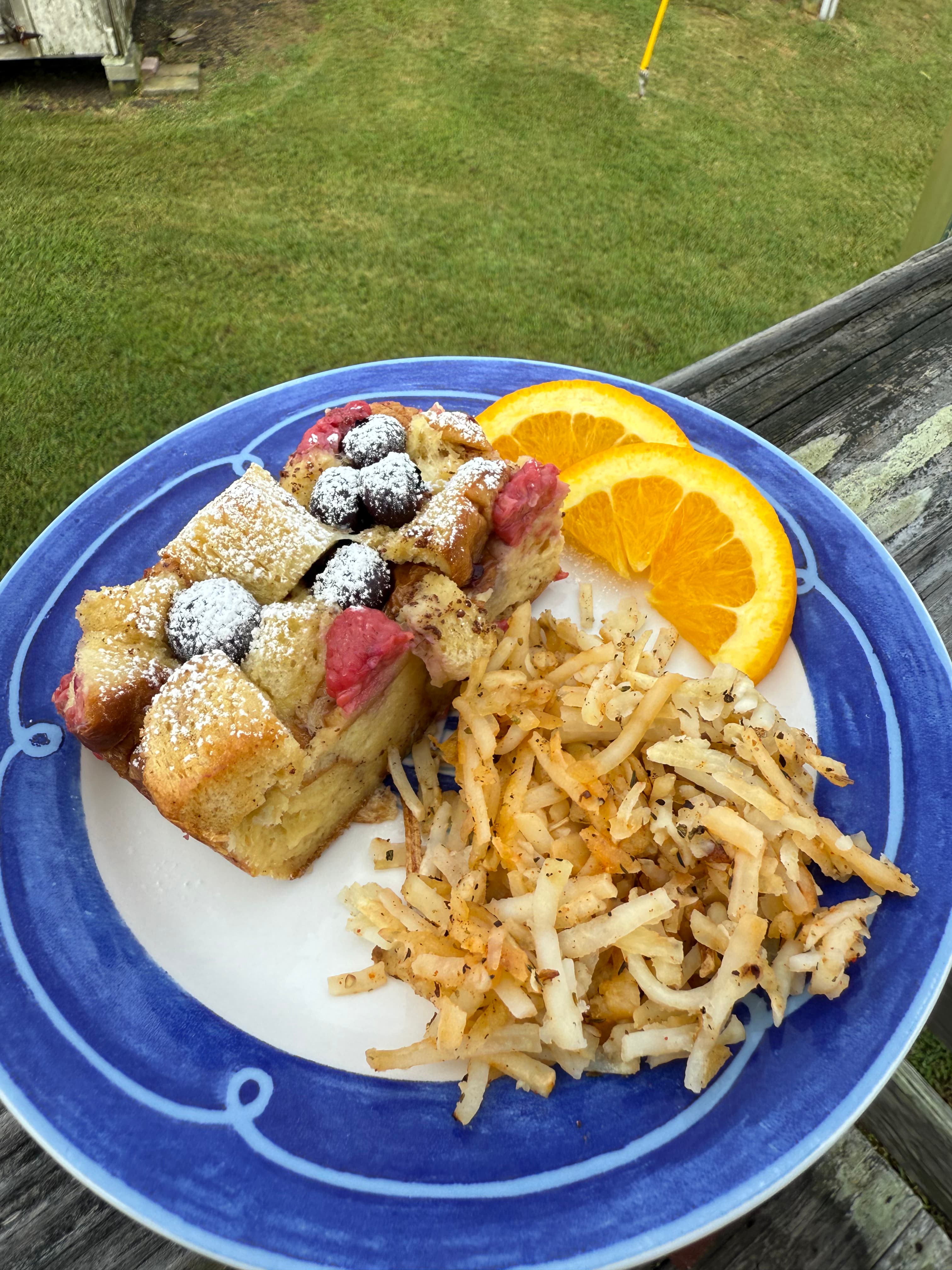 A plate of bread pudding topped with berries, accompanied by hash browns and orange slices.