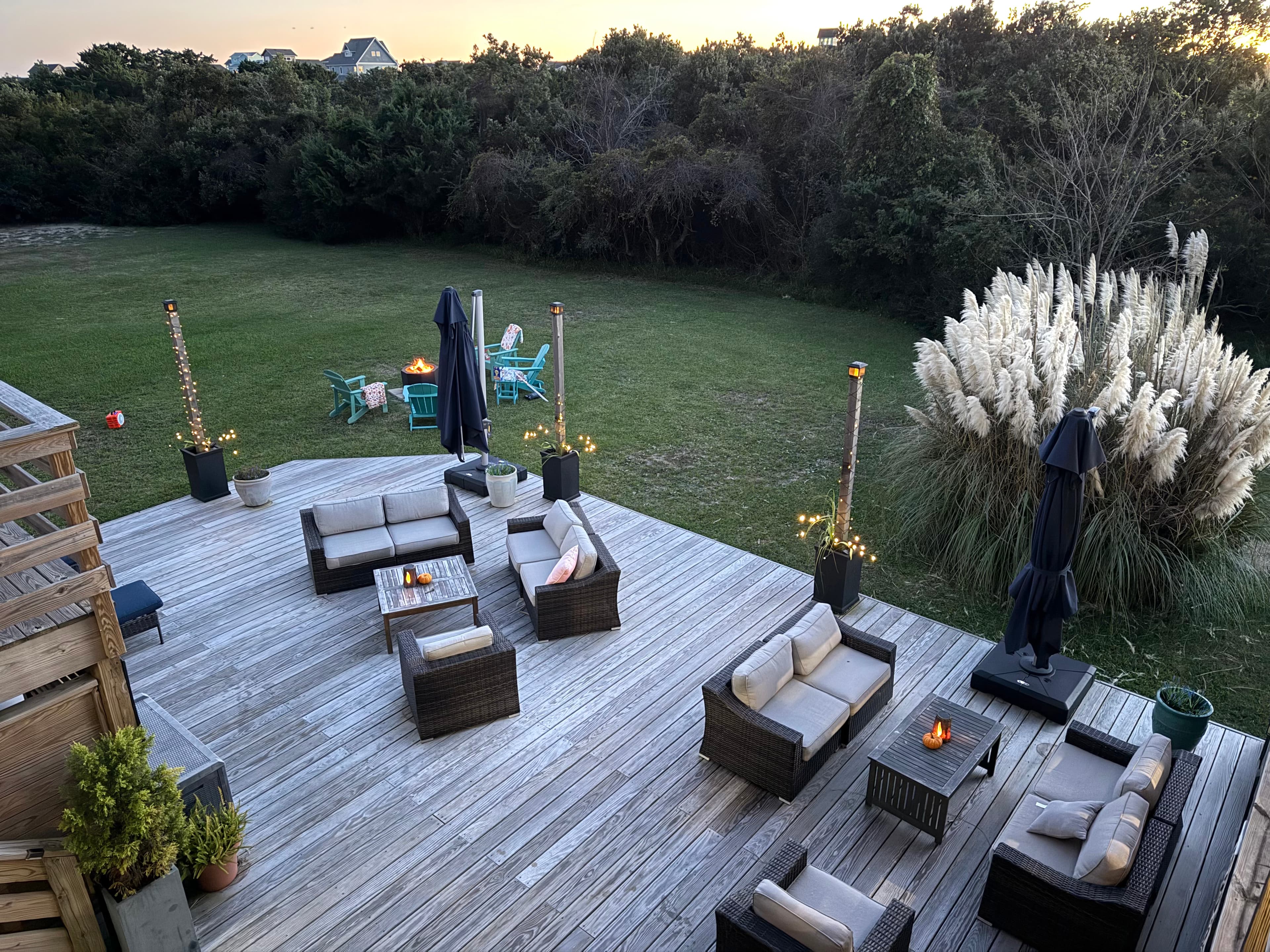 A wooden deck features outdoor seating and fire pits, with a grassy yard and trees in the background.