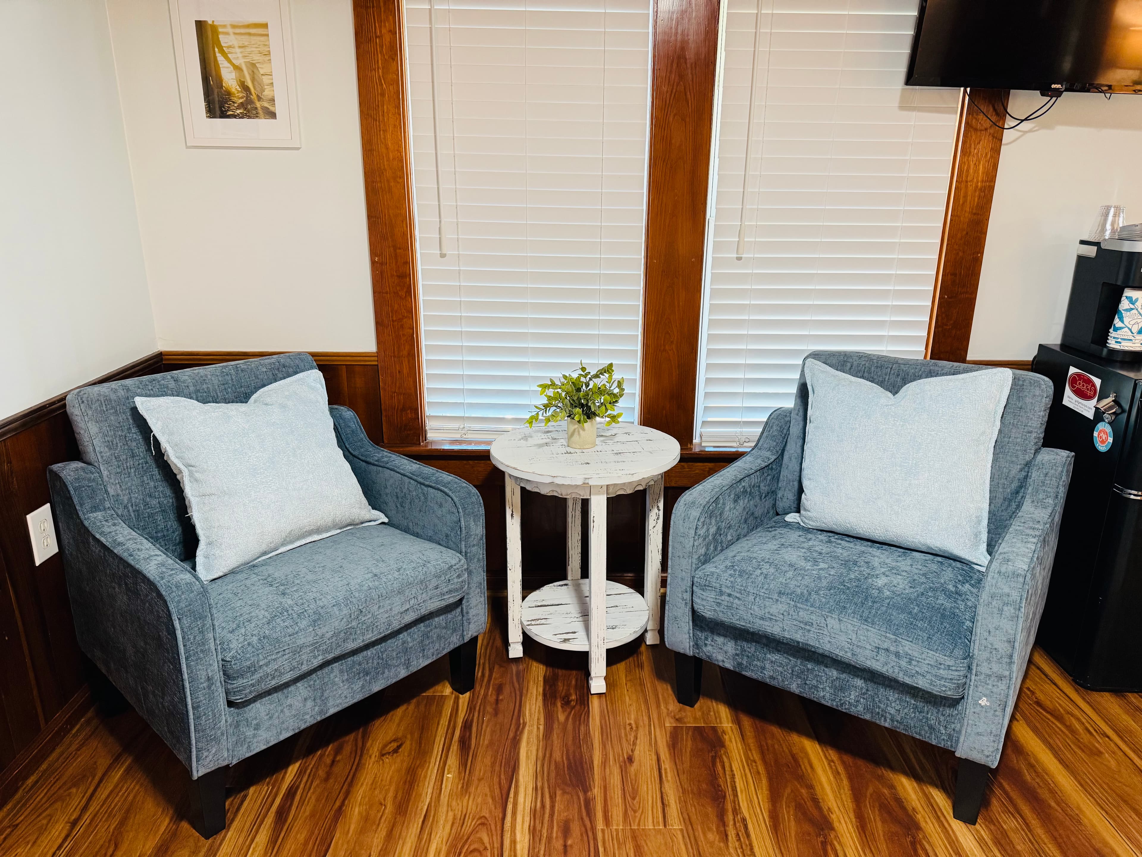 Two gray upholstered chairs with pillows flanking a small white side table with a potted plant, positioned near a window.