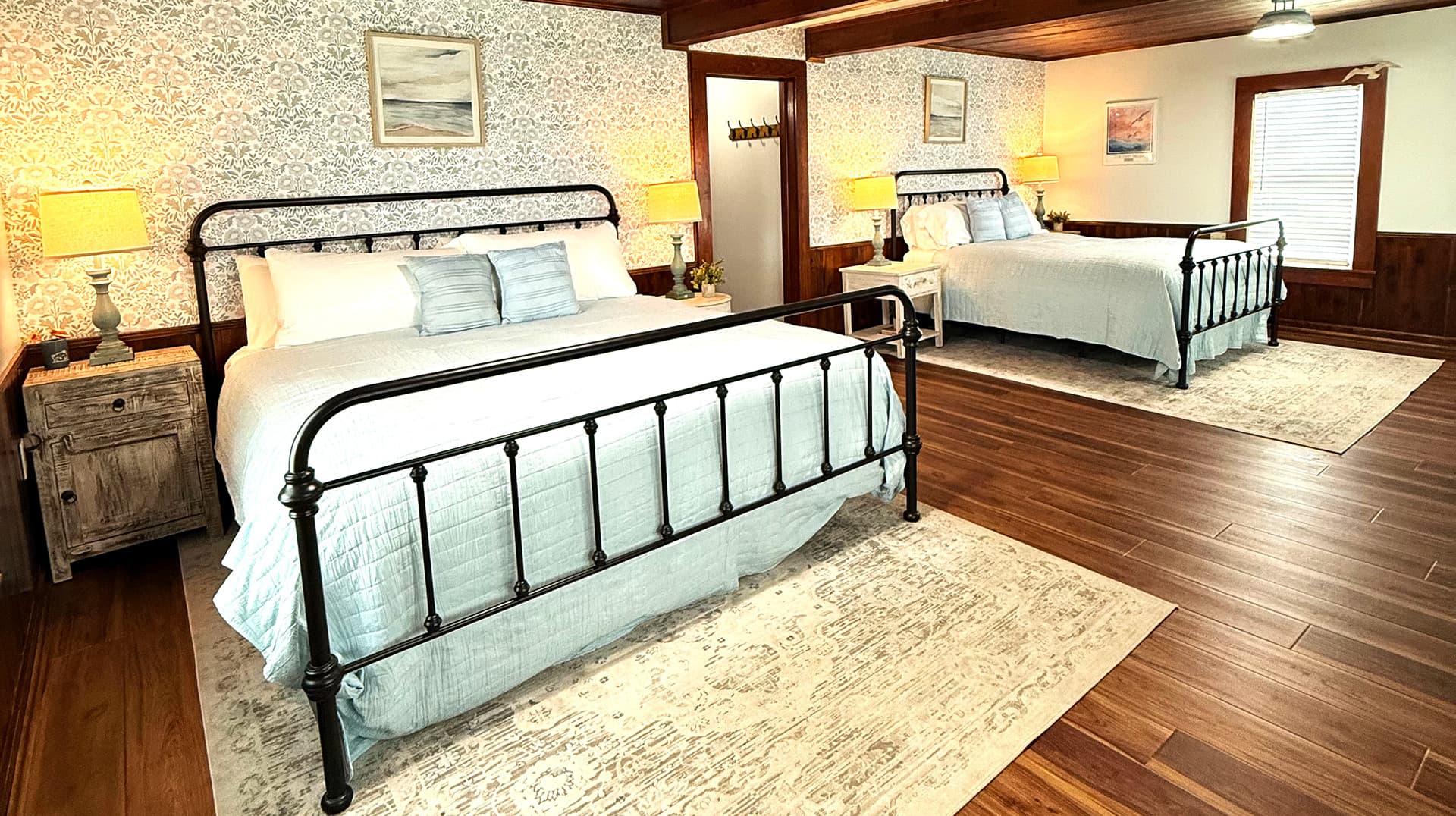 A spacious bedroom featuring two metal-framed beds with light blue bedding and warm wooden accents.