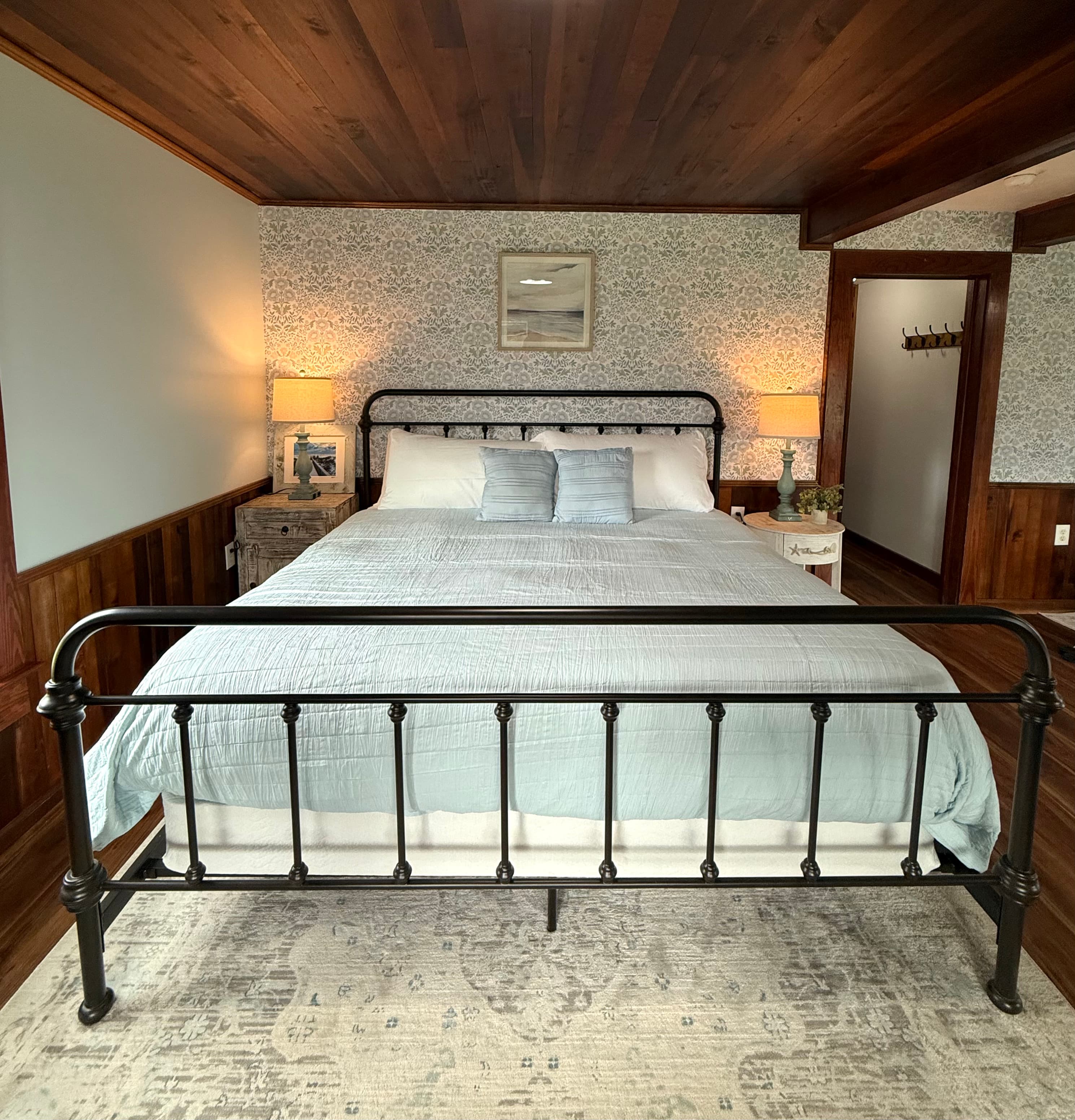 A cozy bedroom featuring a black metal bed frame, light blue bedding, lamps on nightstands, and floral wallpaper.