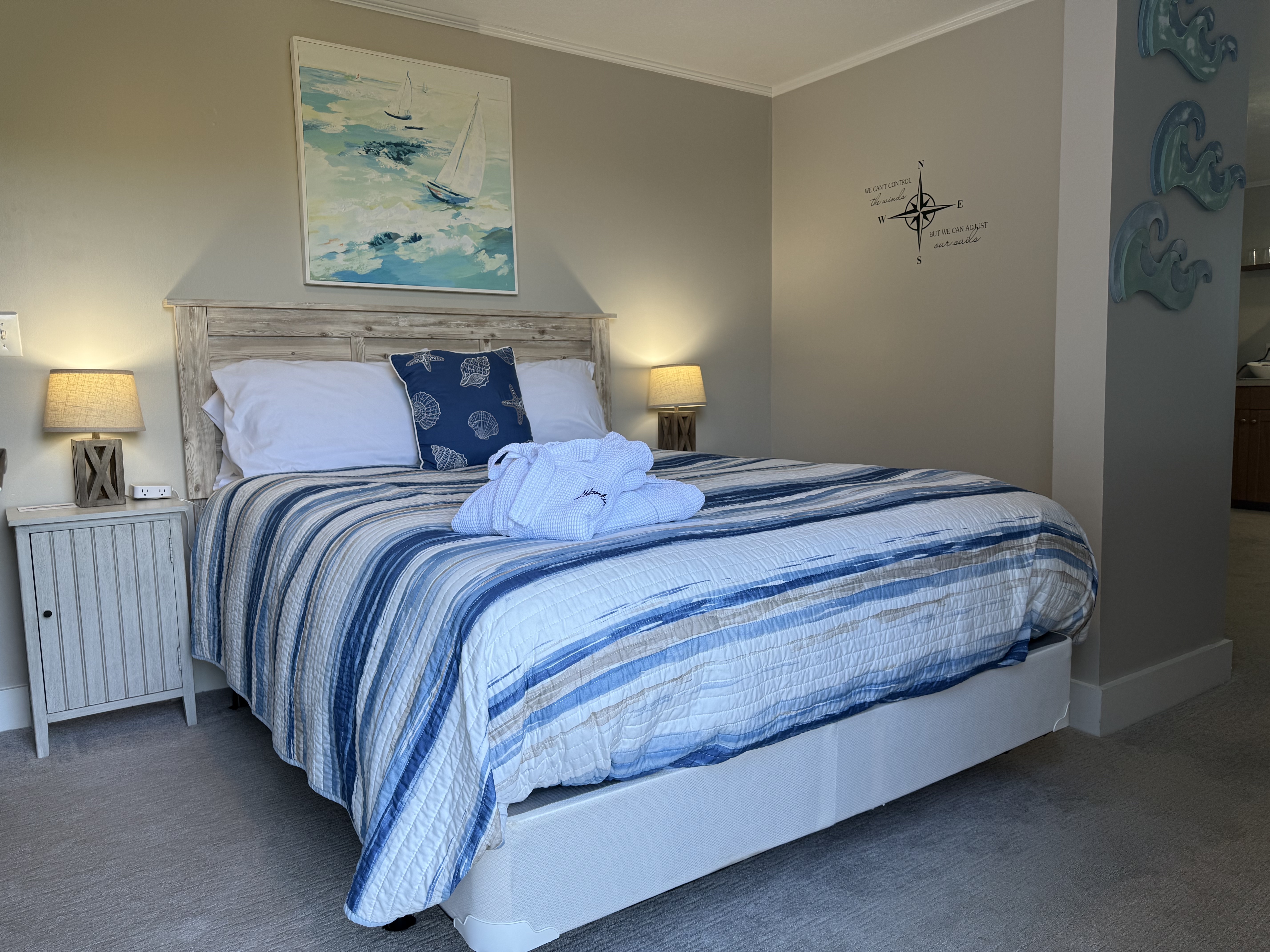 A spacious bedroom at The Atlantic Inn featuring a large bed with blue and white striped bedding, folded white bathrobes, and coastal wall art, with a glimpse into an adjoining living area and kitchenette.
