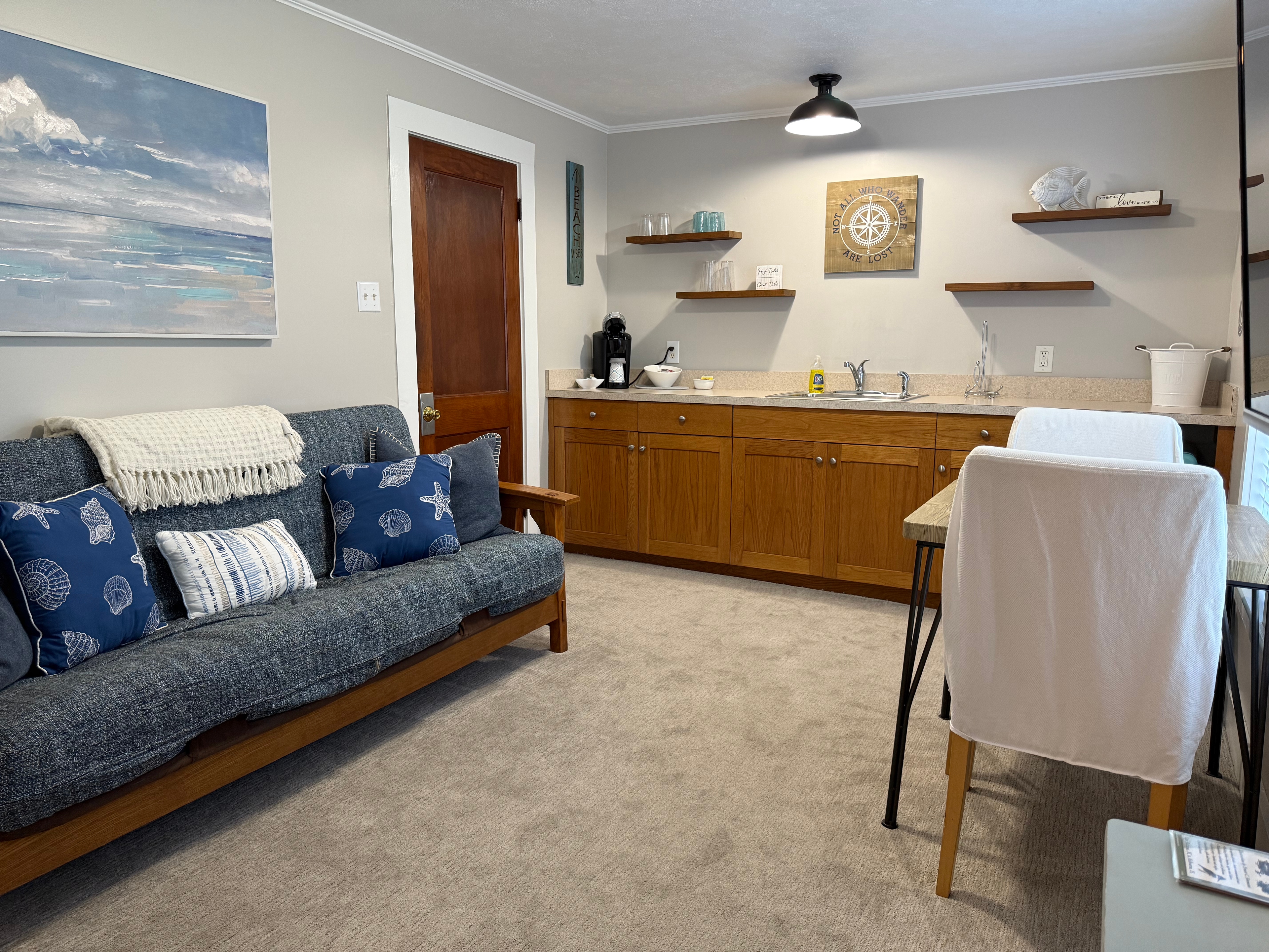 A cozy living area and kitchenette featuring a blue futon with nautical pillows, a wooden wet bar with floating shelves, and a small dining table with white-covered chairs.