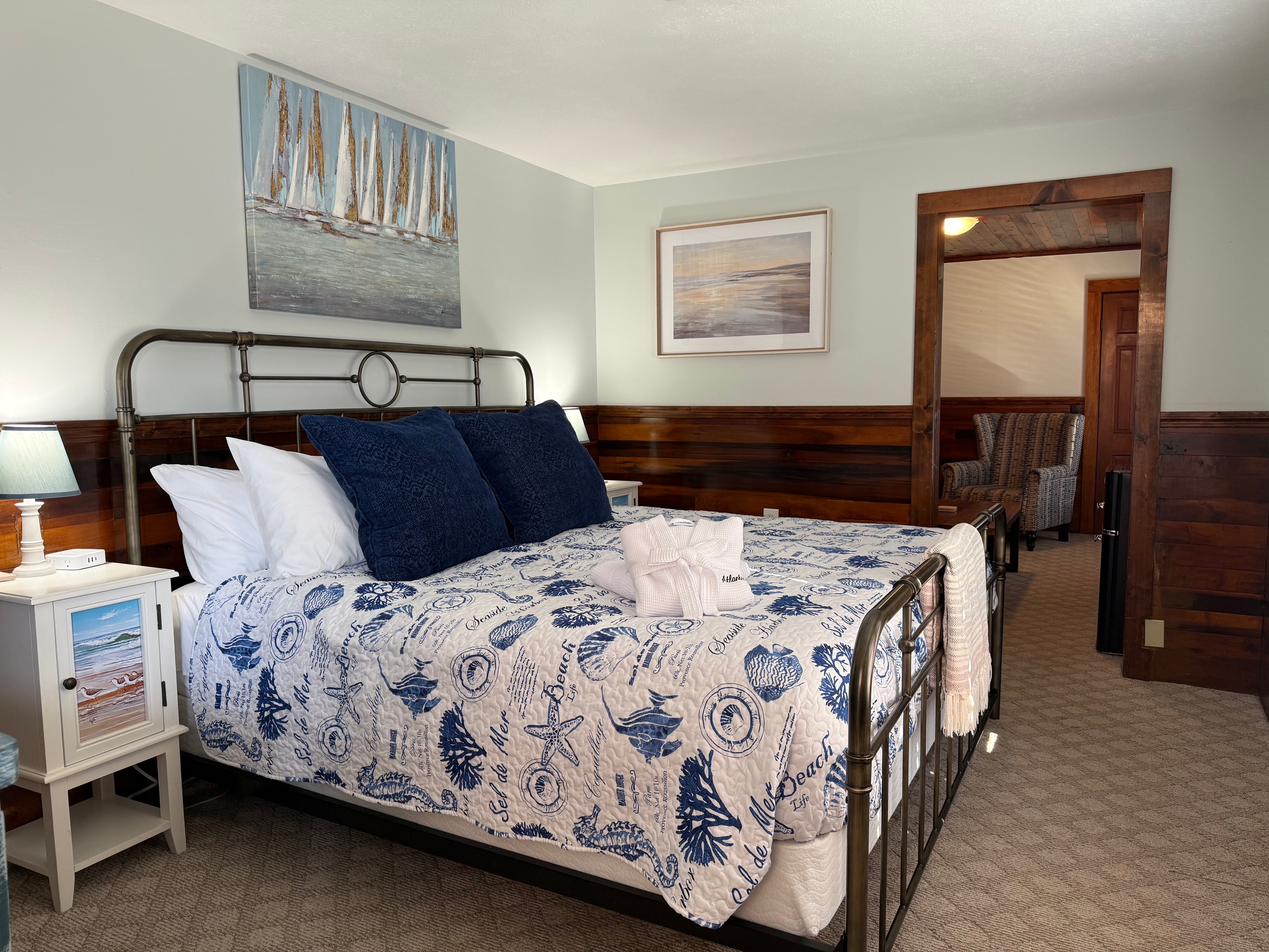 A rustic bedroom at The Atlantic Inn featuring a large bed with nautical seashell-patterned bedding and an iron headboard, set against walls with original dark wood paneling and coastal artwork.