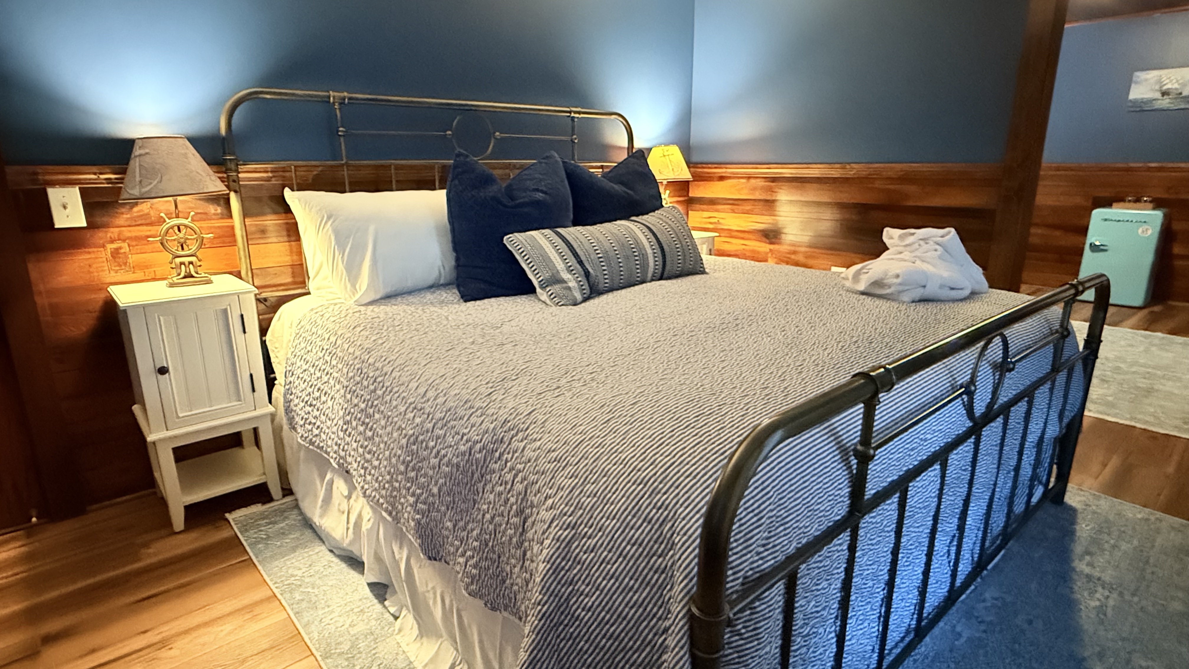 A rustic bedroom at The Atlantic Inn featuring a large bed with nautical seashell-patterned bedding and an iron headboard, set against walls with original dark wood paneling and coastal artwork.