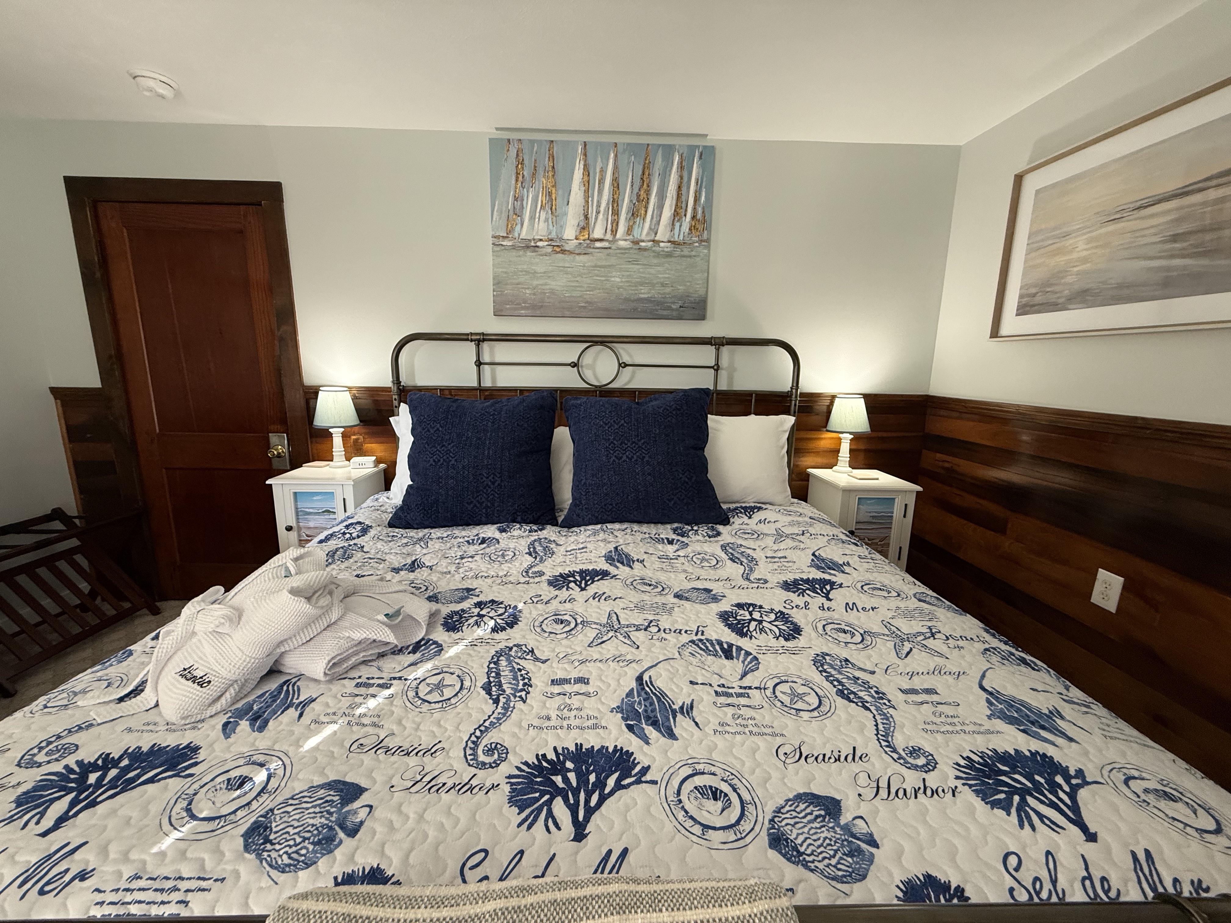 A nautical-themed bedroom at The Atlantic Inn featuring a large bed with blue and white seashell-patterned bedding and an iron headboard. The room is accented with original dark wood wainscoting, coastal artwork, and white bedside tables with lamps.