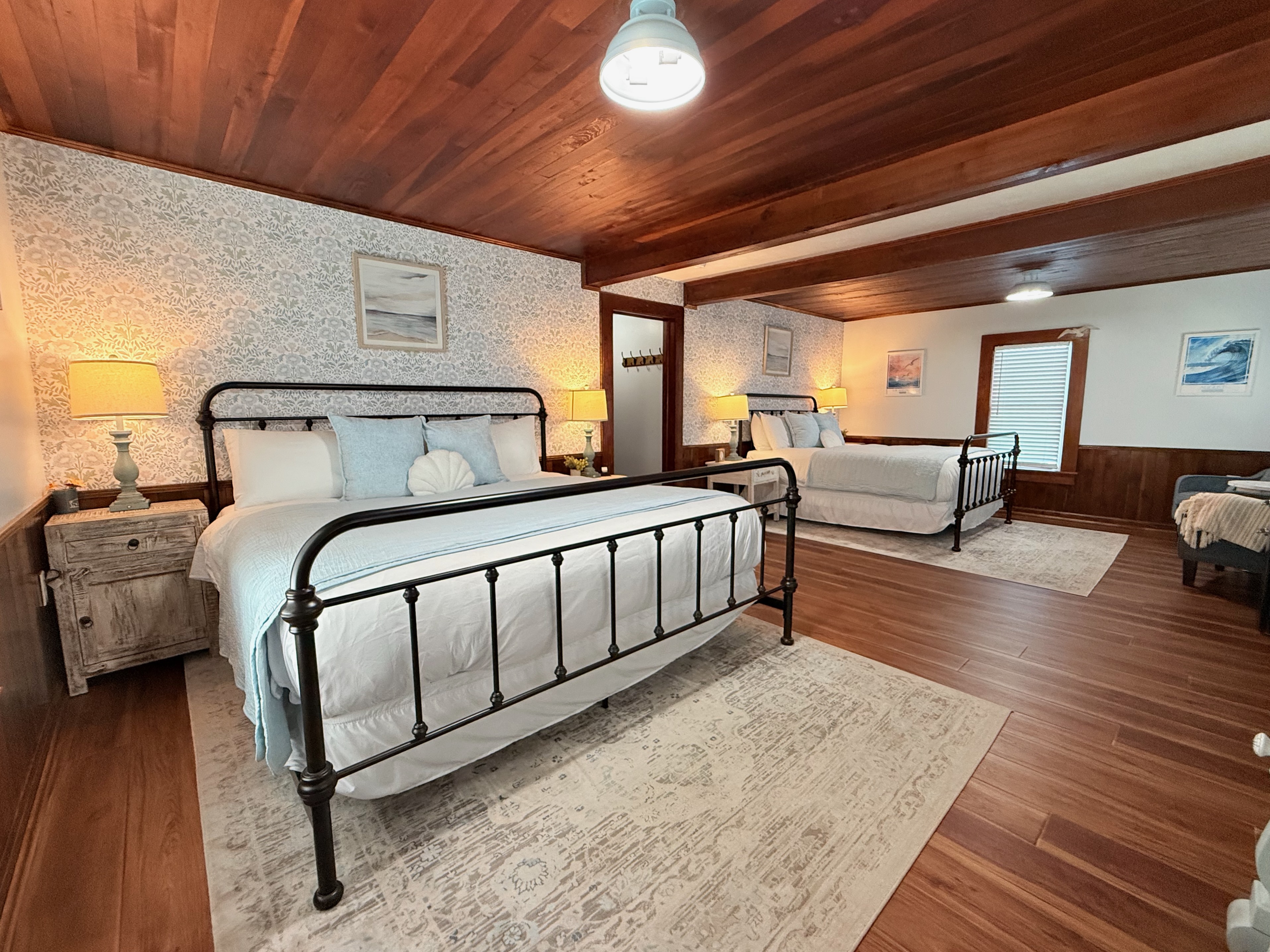 A large, newly remodeled suite featuring a king and a queen bed with iron frames and light blue bedding. The room showcases polished wood floors and a stunning original wood ceiling, accented by coastal wallpaper and warm bedside lighting.