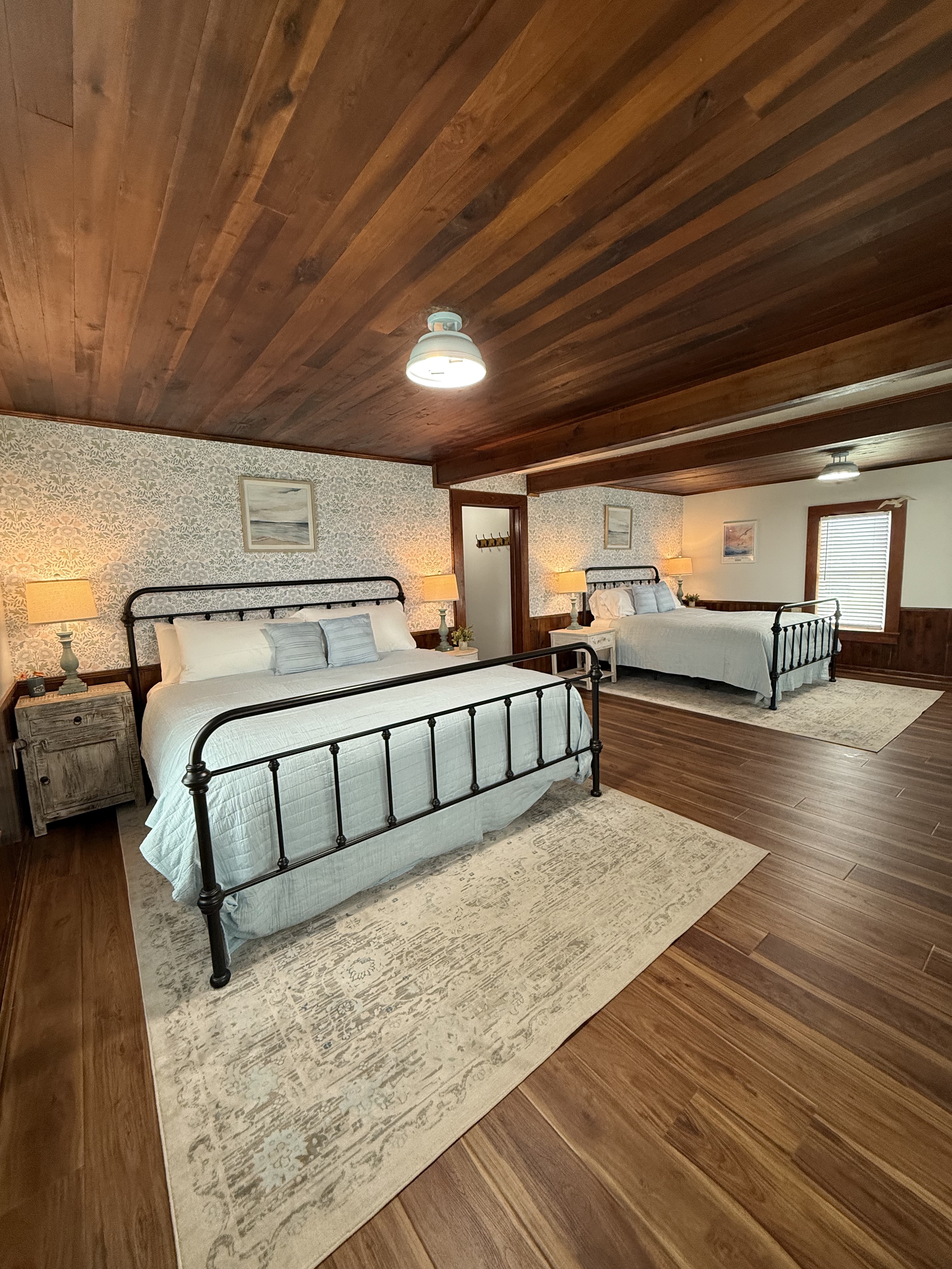A large, newly remodeled suite featuring a king and a queen bed with iron frames and light blue bedding. The room showcases polished wood floors and a stunning original wood ceiling, accented by coastal wallpaper and warm bedside lighting.