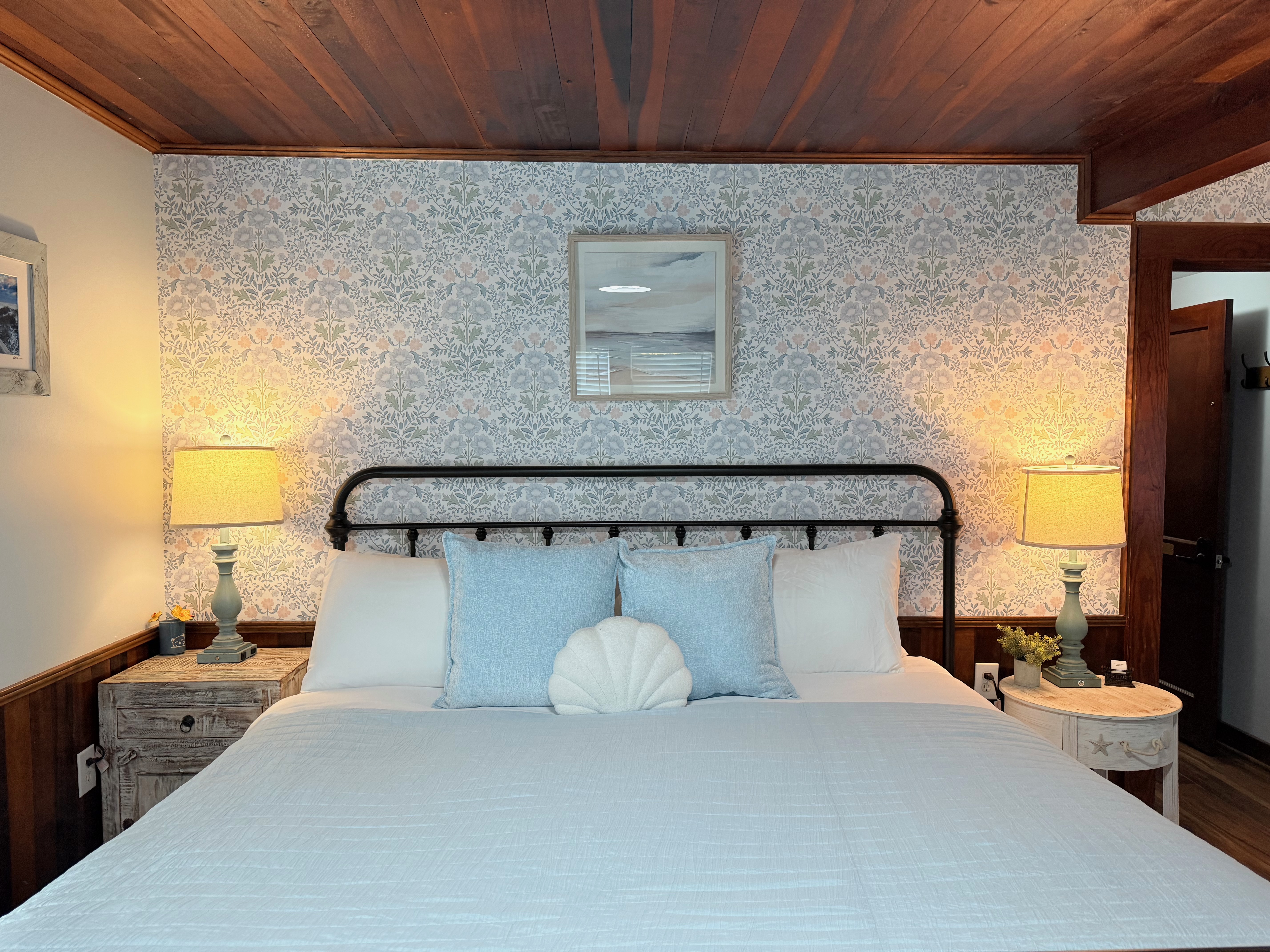 A large, newly remodeled suite featuring a king bed and a queen bed with iron frames and light blue bedding. The room showcases polished wood floors and a stunning original wood ceiling, accented by coastal wallpaper and warm bedside lighting.