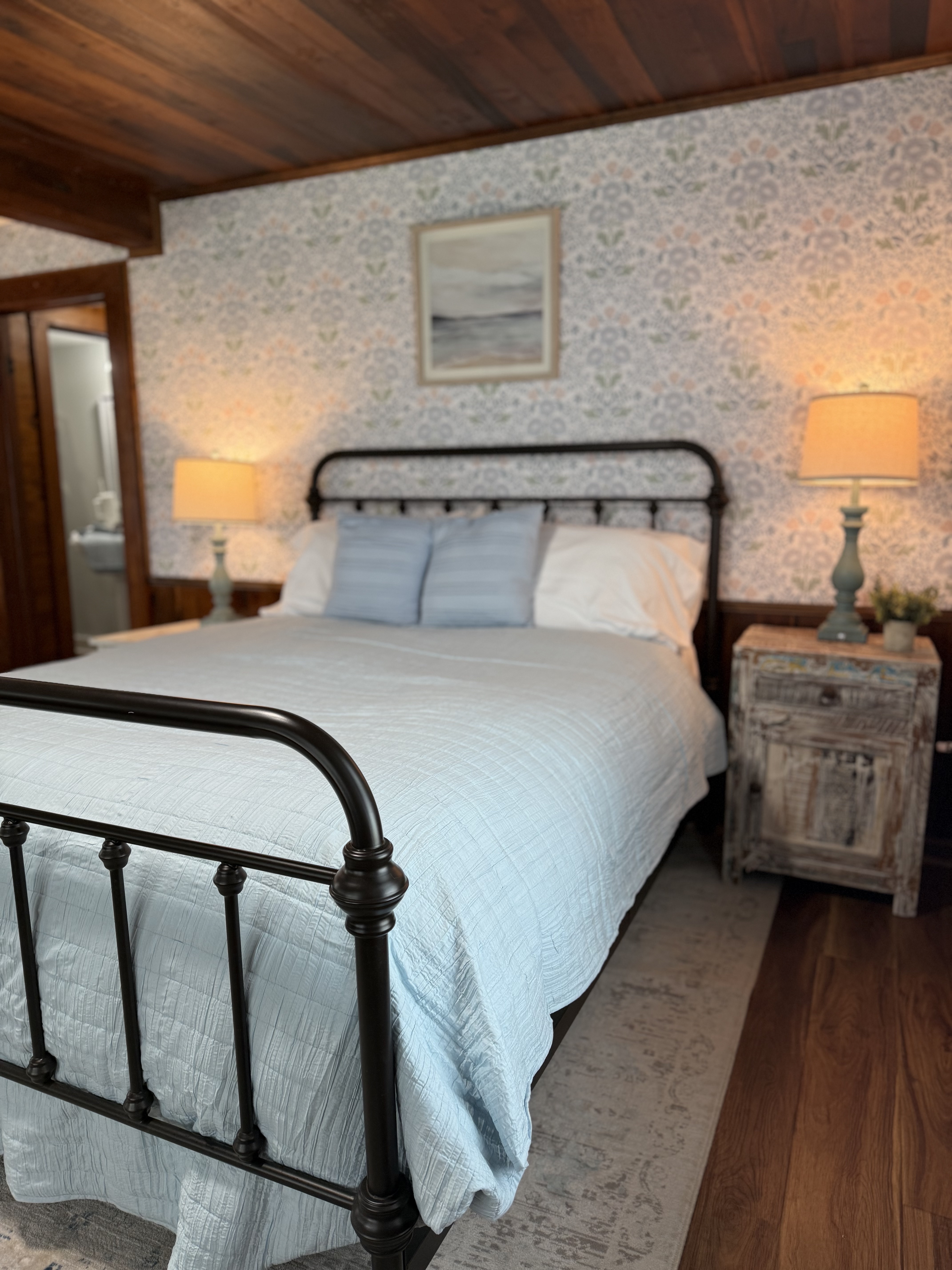A detail view of a bedroom in a newly remodeled suite featuring a queen-size bed with an iron frame and light blue bedding. The room is decorated with coastal-patterned wallpaper, a rustic wooden nightstand with a lamp, and original dark wood ceilings and flooring.