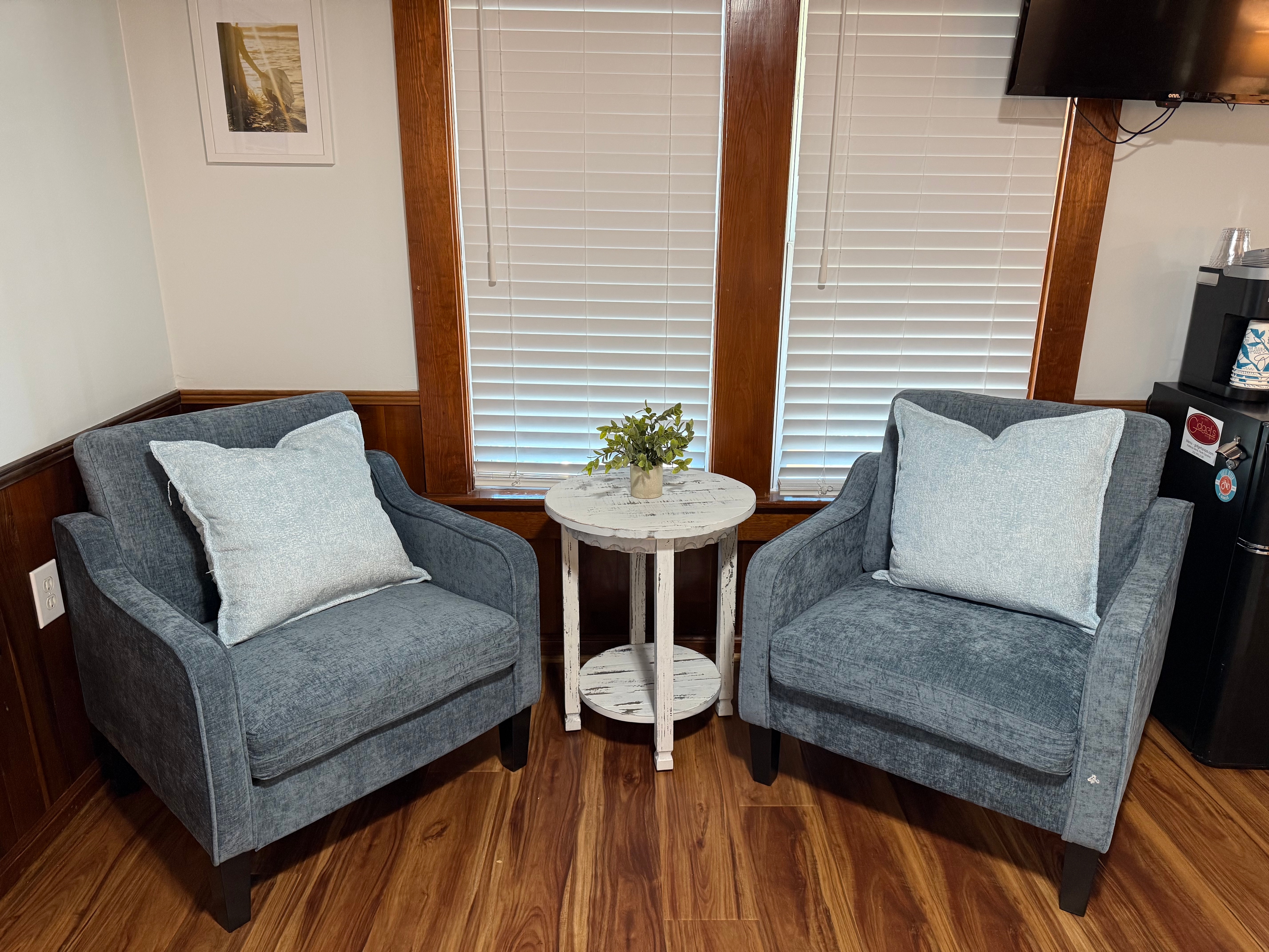 A cozy reading nook featuring two denim-blue upholstered armchairs with light blue pillows, centered around a small white round side table. The space is accented by warm wood flooring, wood-trimmed windows with white blinds, and a wall-mounted TV.