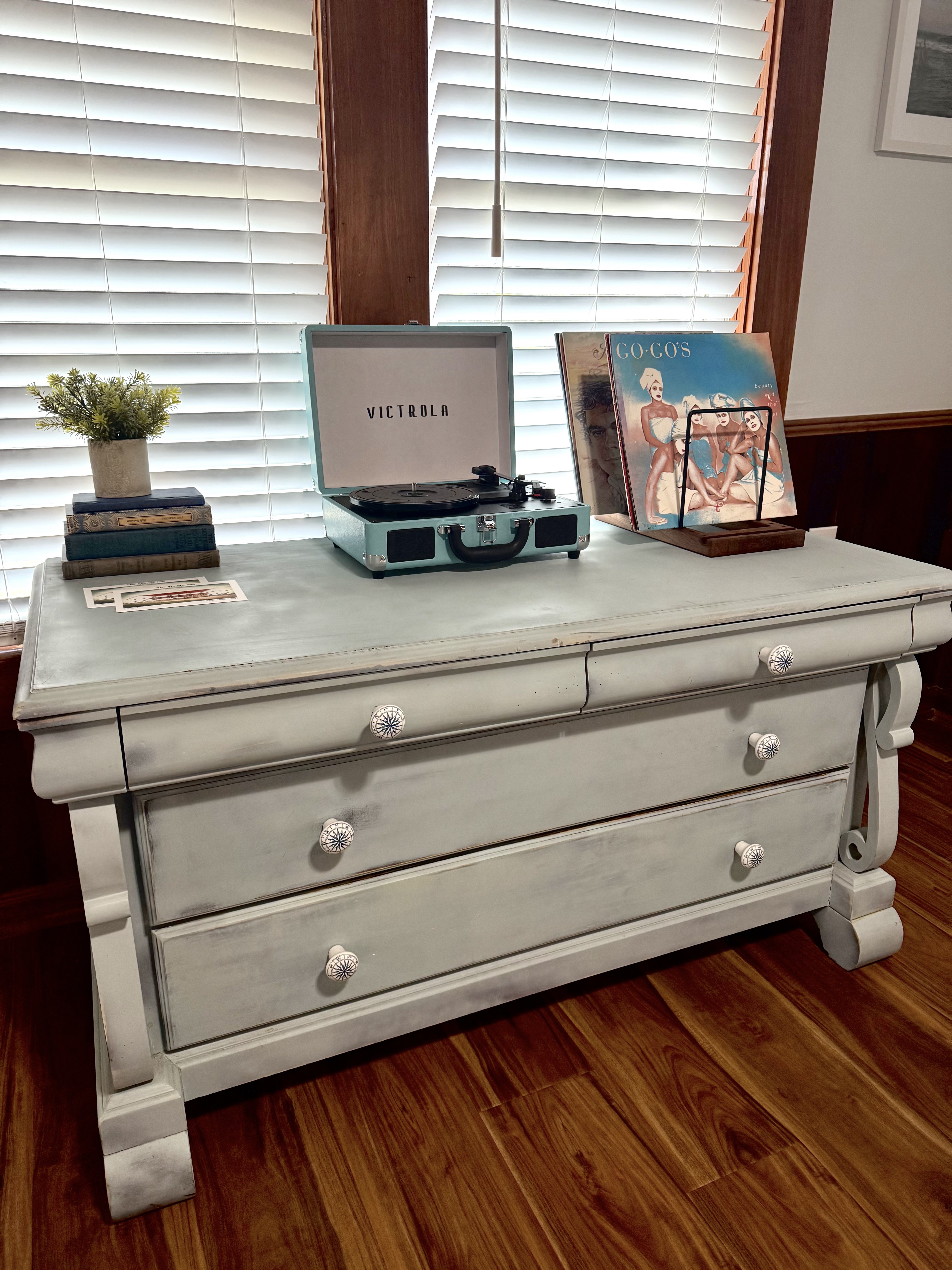 A light blue vintage-style suitcase record player sitting on a distressed white wooden dresser. Next to the player is a stand holding vinyl records, all positioned in front of a window with white blinds and dark wood trim.