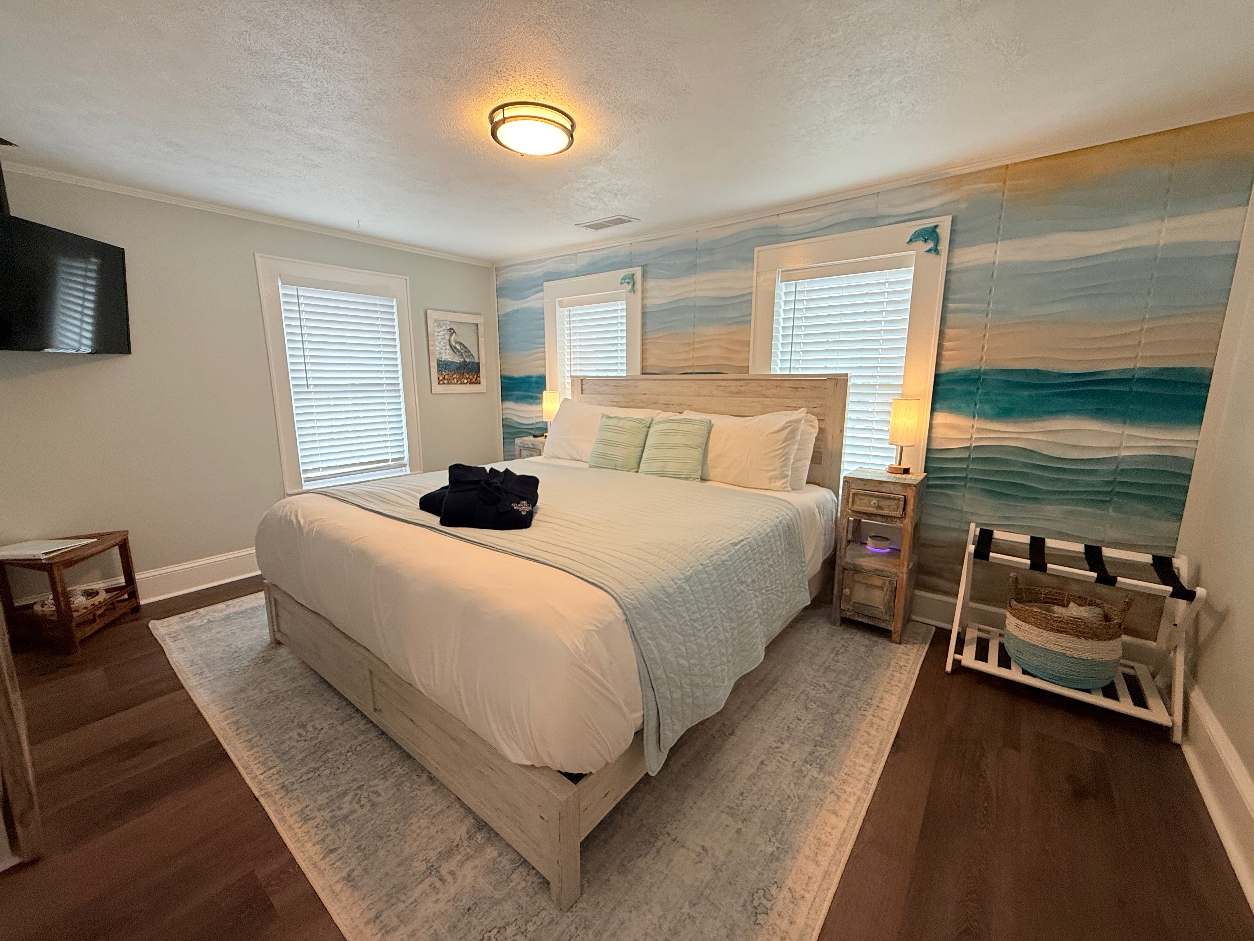 A brightly lit bedroom featuring a large bed with a light-colored wood headboard and white linens. The wall behind the bed is covered in coastal-themed wallpaper with blue, tan, and white horizontal waves, and two windows with white blinds flank the bed.