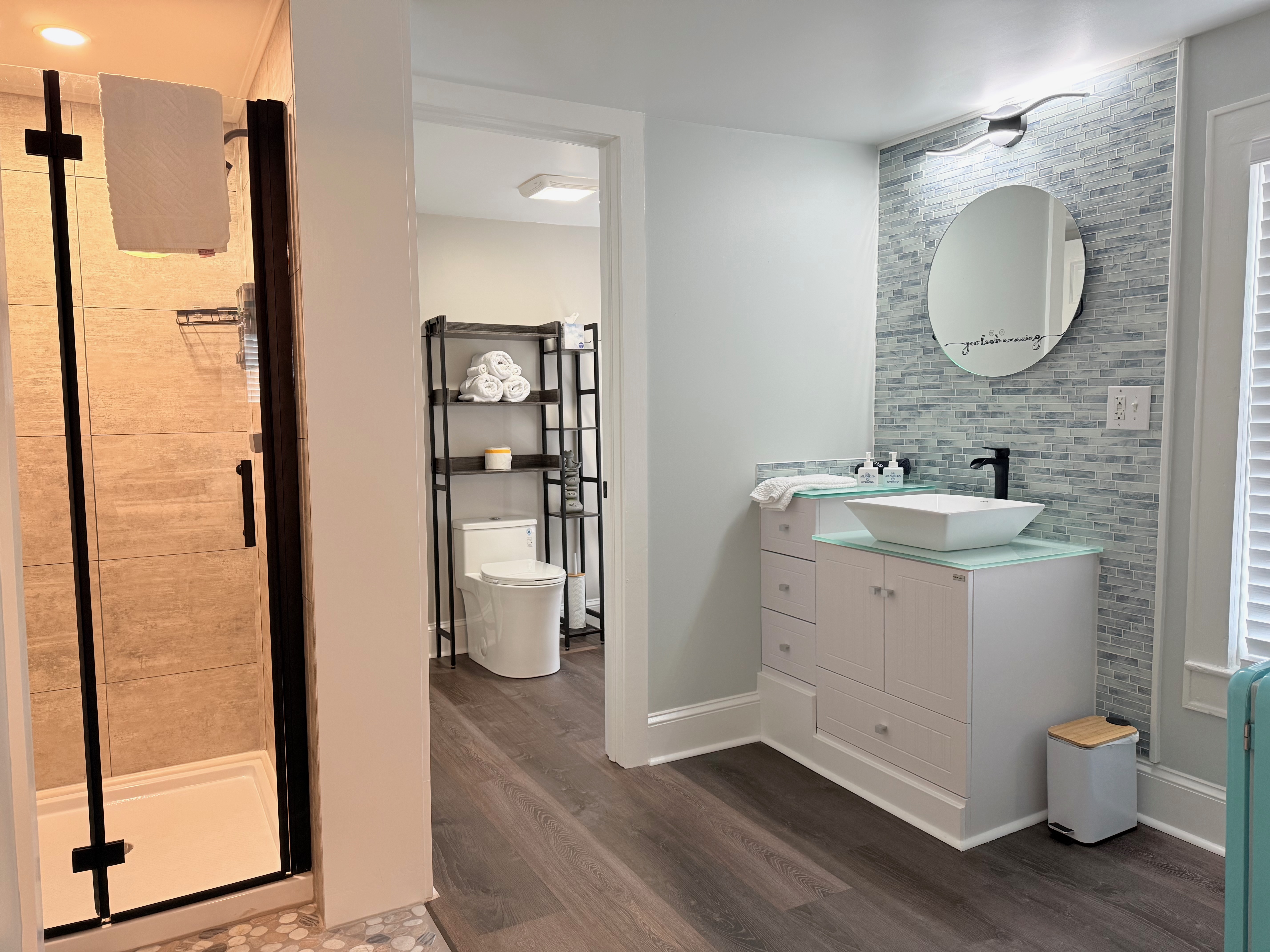 A modern walk-in shower featuring light gray wood-look tile walls, a white shower base, and a pebble-style floor. The shower is equipped with a black rainfall shower head and a glass door with a black frame.