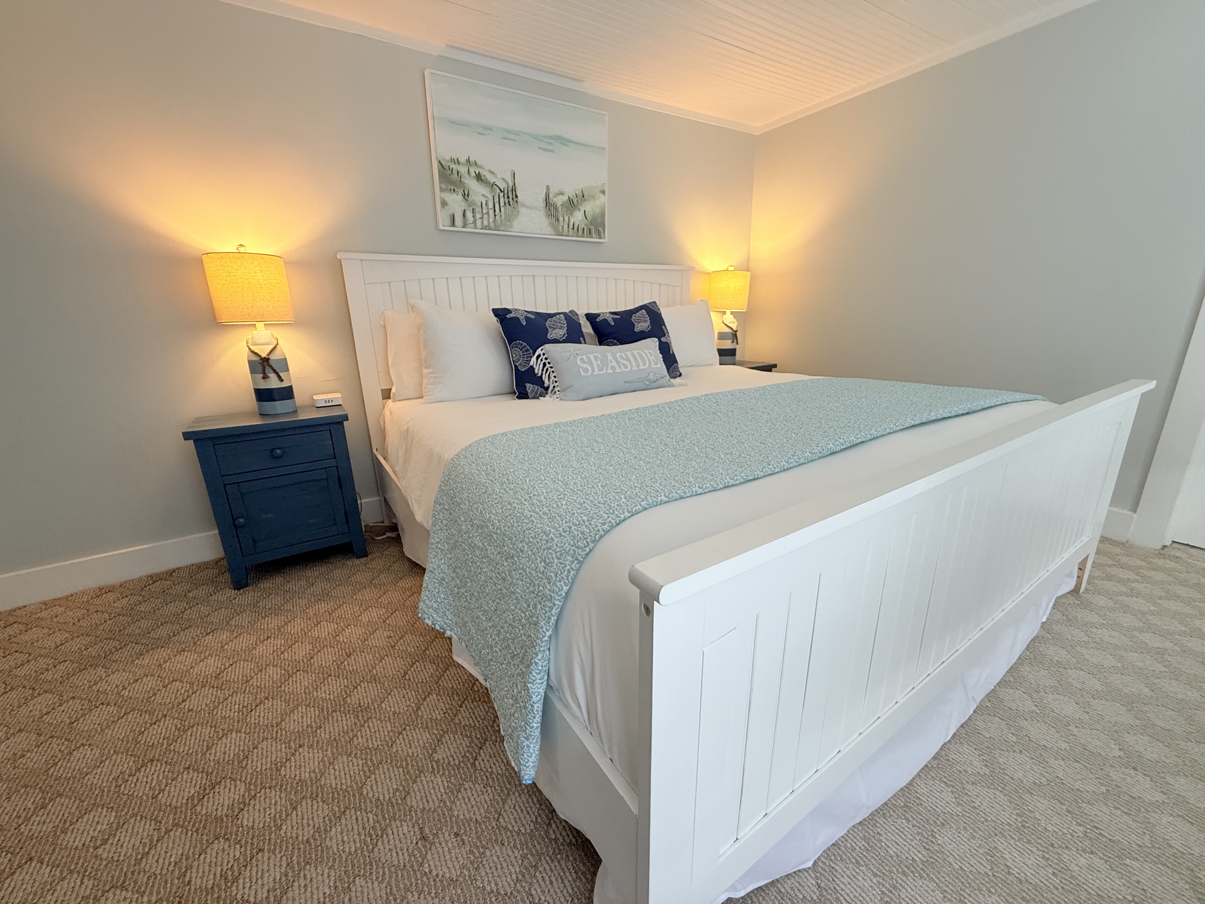 A guest room featuring a large white wooden bed frame with a navy and white striped comforter. The room includes a blue bedside table with a lamp, light gray walls, and a coastal landscape painting hanging above the headboard.