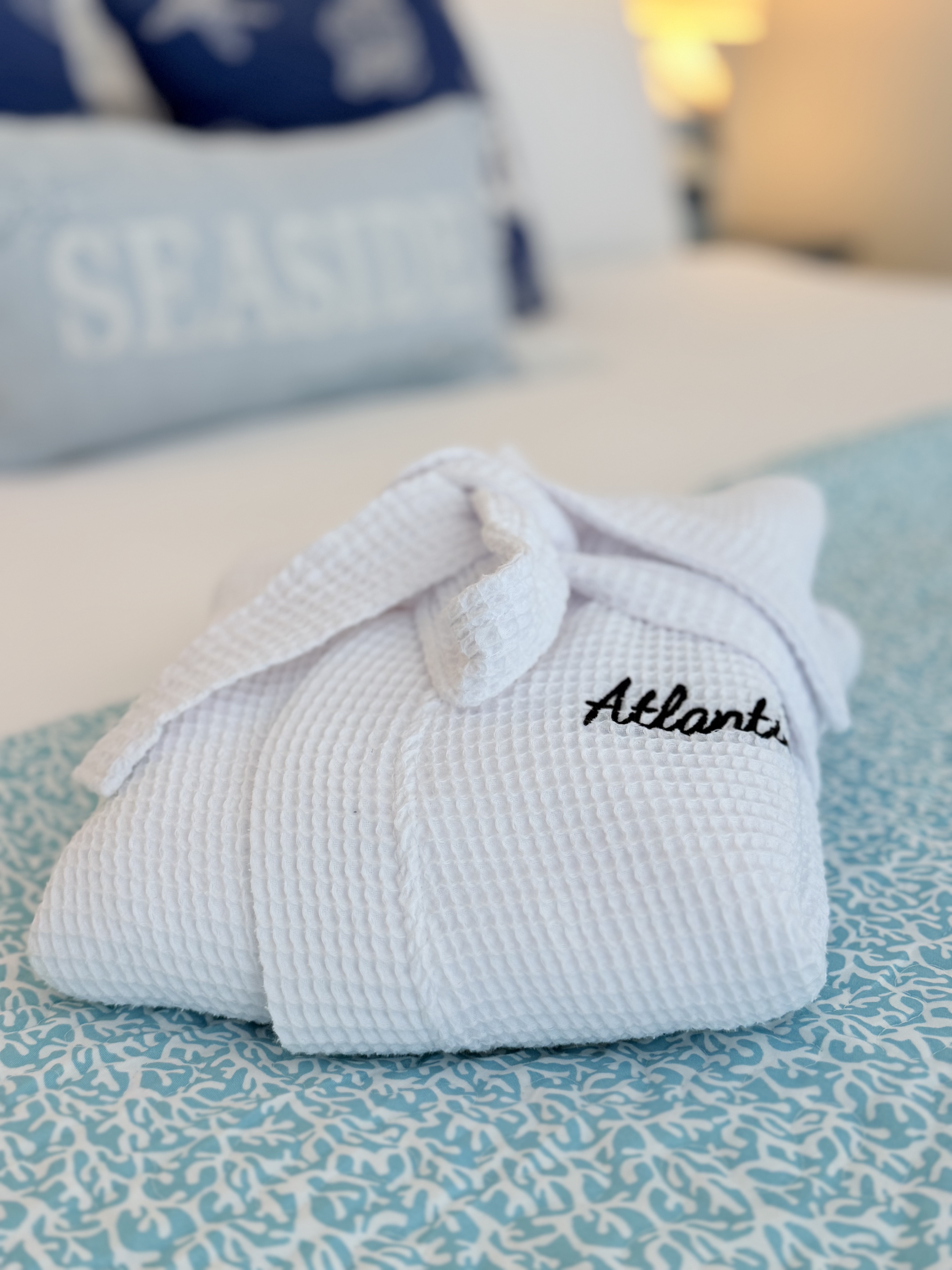 A brightly lit bedroom featuring a large bed with a white wooden headboard and a navy and white striped comforter. The room includes blue starfish accent pillows, light gray walls, and a coastal painting of sand dunes hanging above the bed.