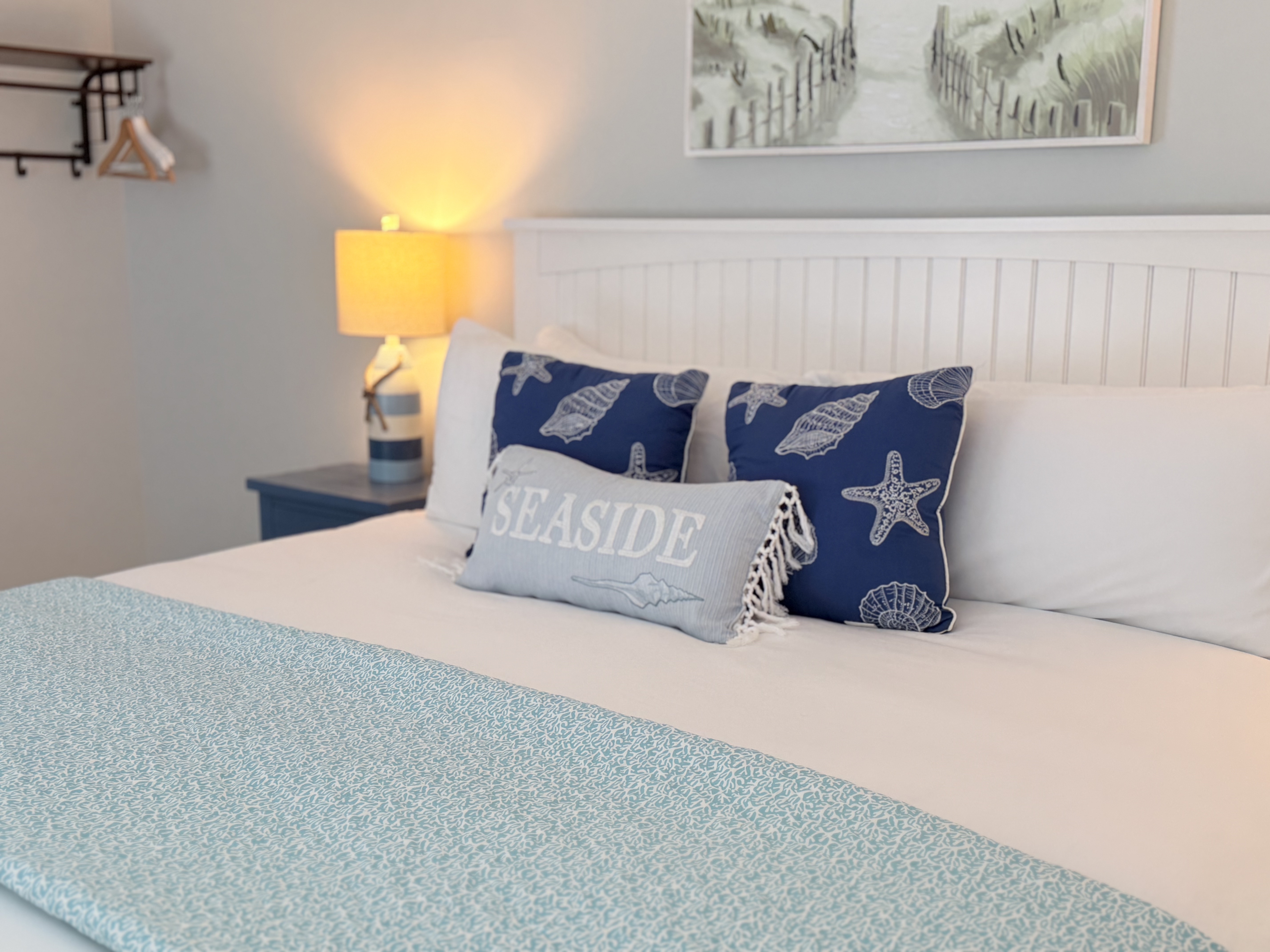 A brightly lit bedroom featuring a large bed with a white wooden headboard and a navy and white striped comforter. The room includes blue starfish accent pillows, light gray walls, and a coastal painting of sand dunes hanging above the bed.