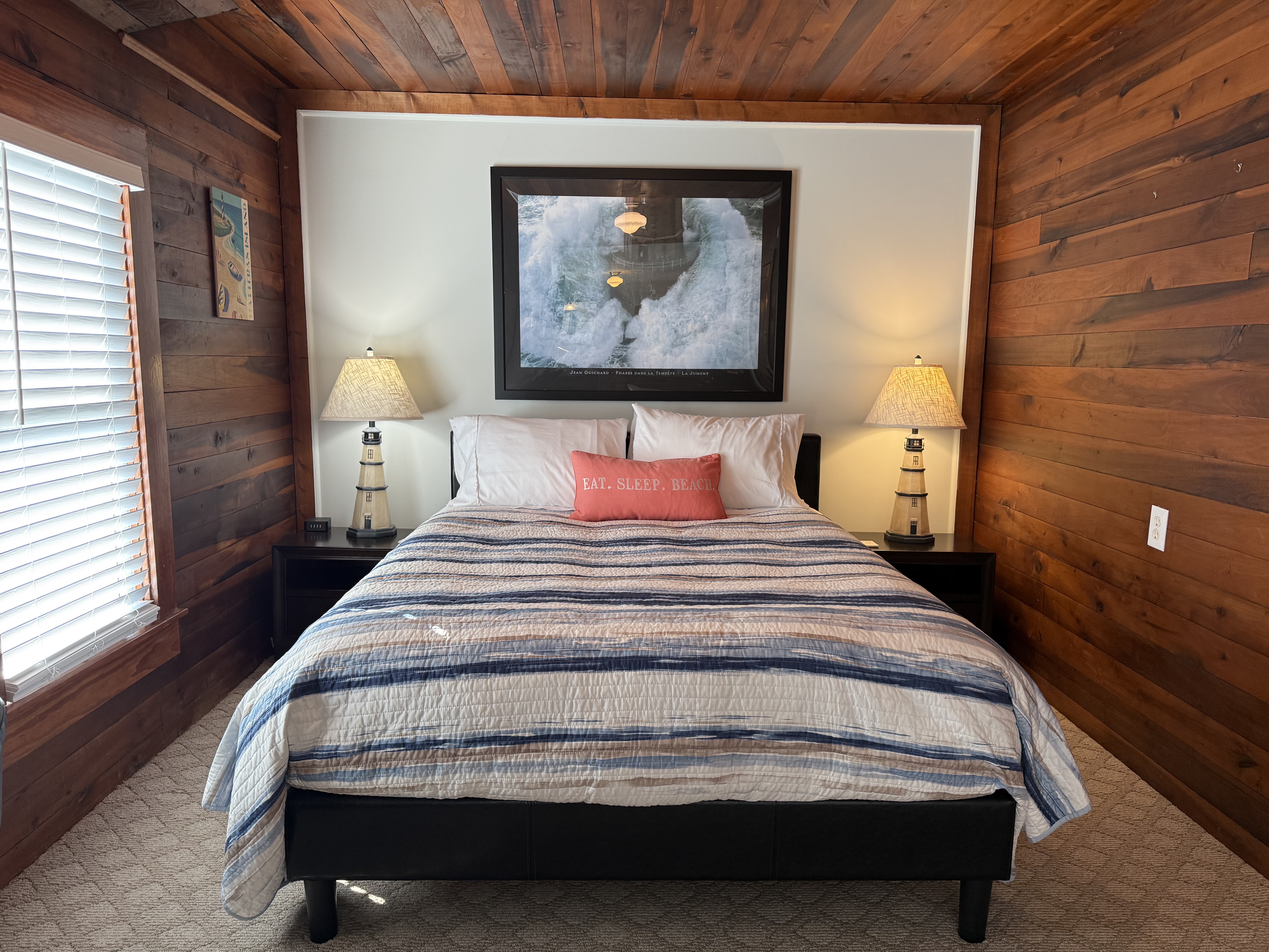 A rustic guest room featuring rich natural wood-paneled walls and ceiling. The bed is styled with a blue and white striped comforter and a coral accent pillow, flanked by two lighthouse-themed bedside lamps. A large framed picture hangs on the white accent wall above the headboard.