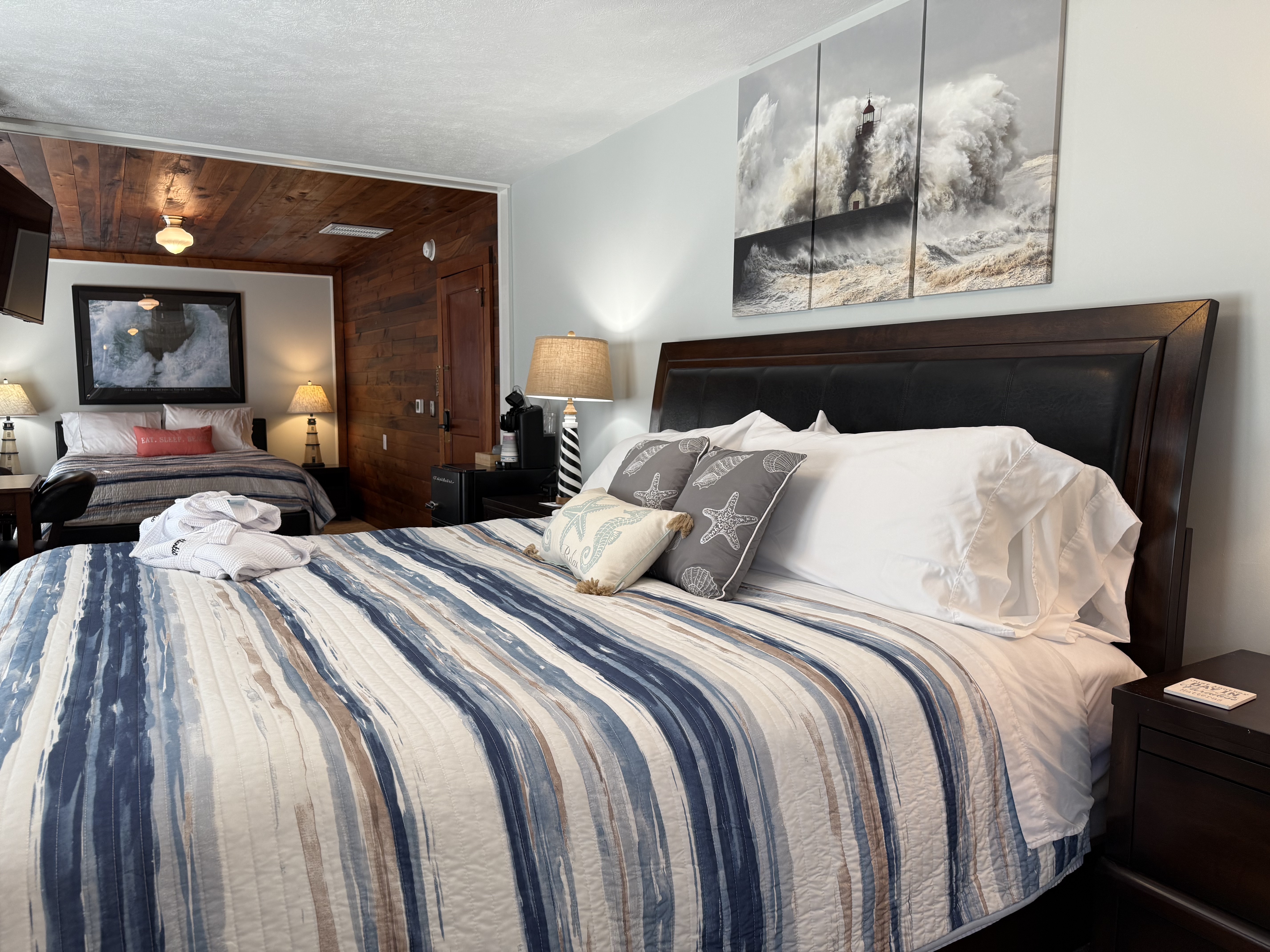 A suite featuring two beds with navy and white striped comforters. The foreground bed has a dark wood headboard and starfish accent pillows, while the background bed is situated in a cozy nook with warm wood-paneled walls and ceiling. A large three-panel canvas of a crashing ocean wave hangs above the main bed.