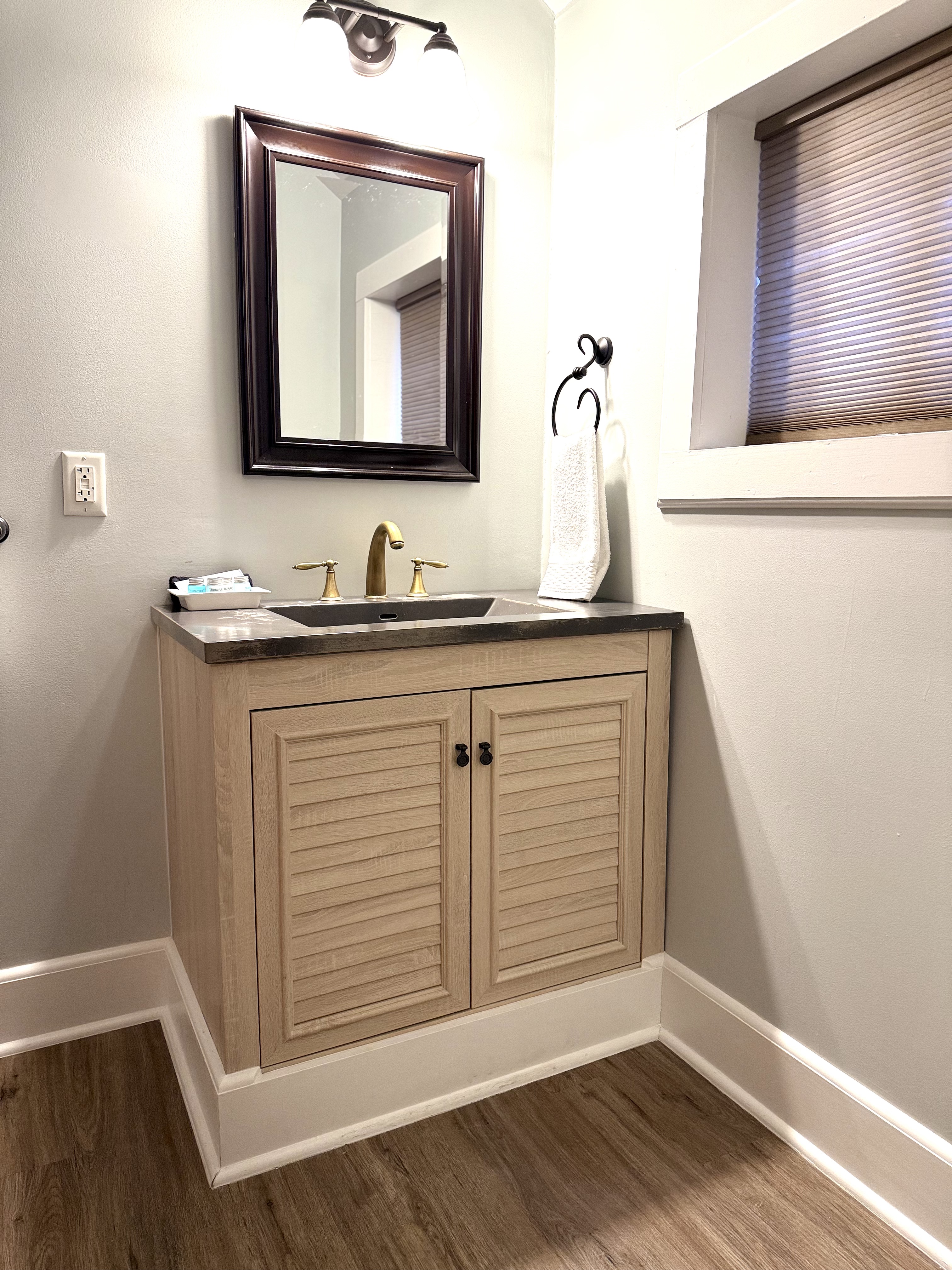 A modern bathroom vanity featuring light wood louvered cabinet doors and a dark countertop with a brass faucet. A dark-framed mirror hangs on the light gray wall above the vanity, and a window with a brown shade is visible to the right.