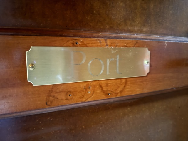 A close-up of a polished brass nameplate engraved with the word "Port" in a serif font, mounted on a rich, dark-toned wooden door.