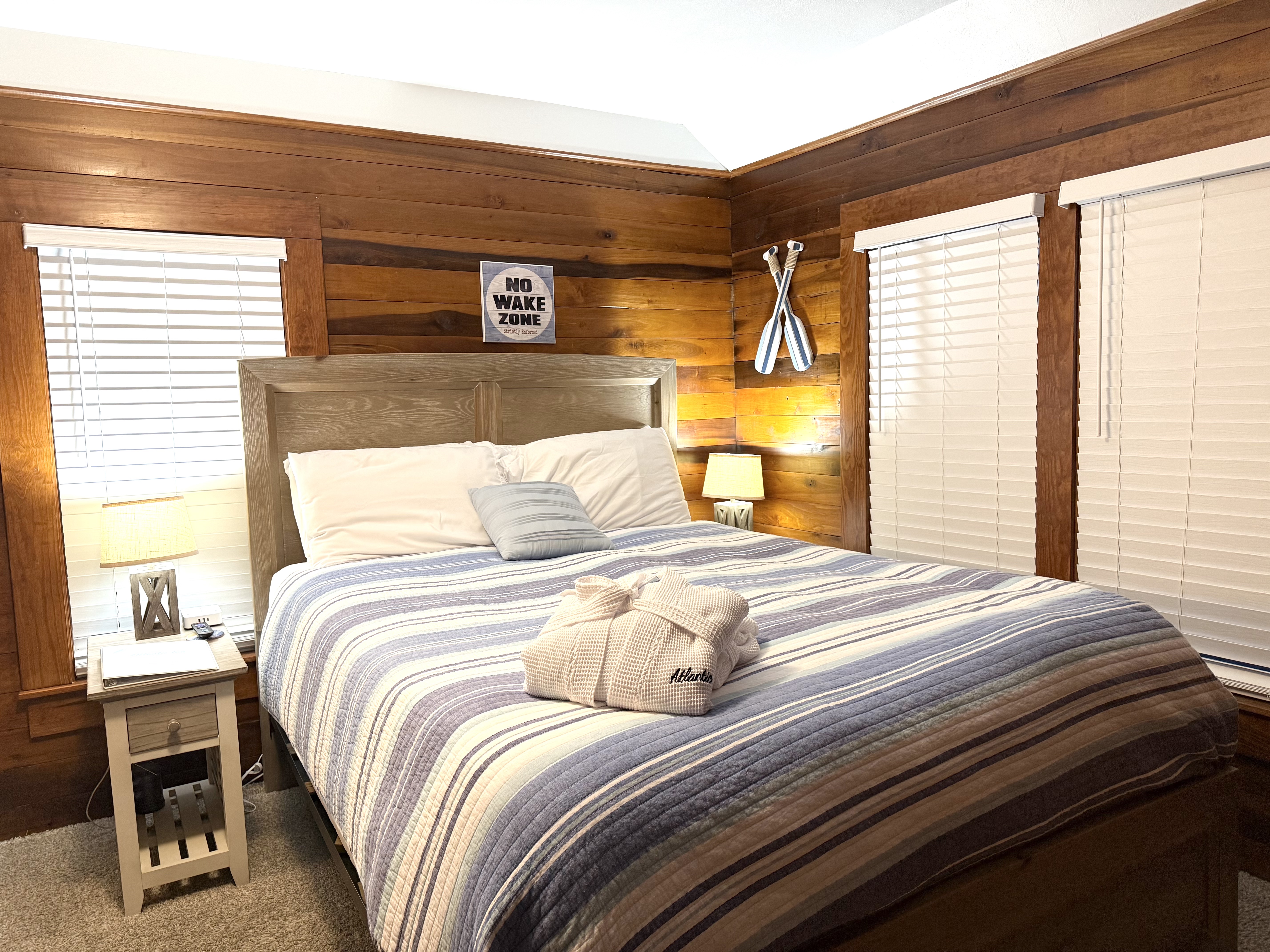 A rustic guest room featuring warm, natural wood-paneled walls and a large bed with a light wood headboard. The bed is styled with a blue and white striped comforter and a white plush robe, flanked by two windows with white blinds and nautical-themed decor, including silver oars mounted on the wall.