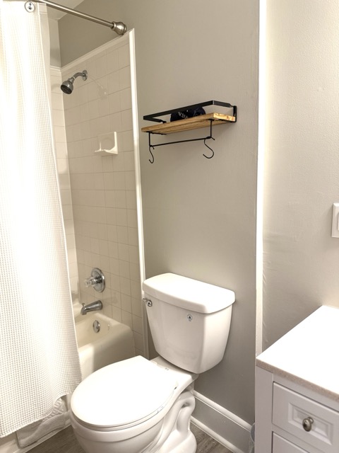 A clean, minimalist guest bathroom featuring a white toilet and a white bathtub with a matching shower curtain. A small wooden shelf with black metal hardware and hooks is mounted on the light gray wall above the toilet.