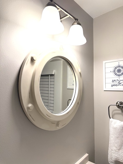 A close-up of a unique bathroom mirror, designed to look like a white circular ship's porthole. The mirror is mounted on a light gray wall beneath a triple-bulb vanity light, with a nautical-themed framed print visible in the background.