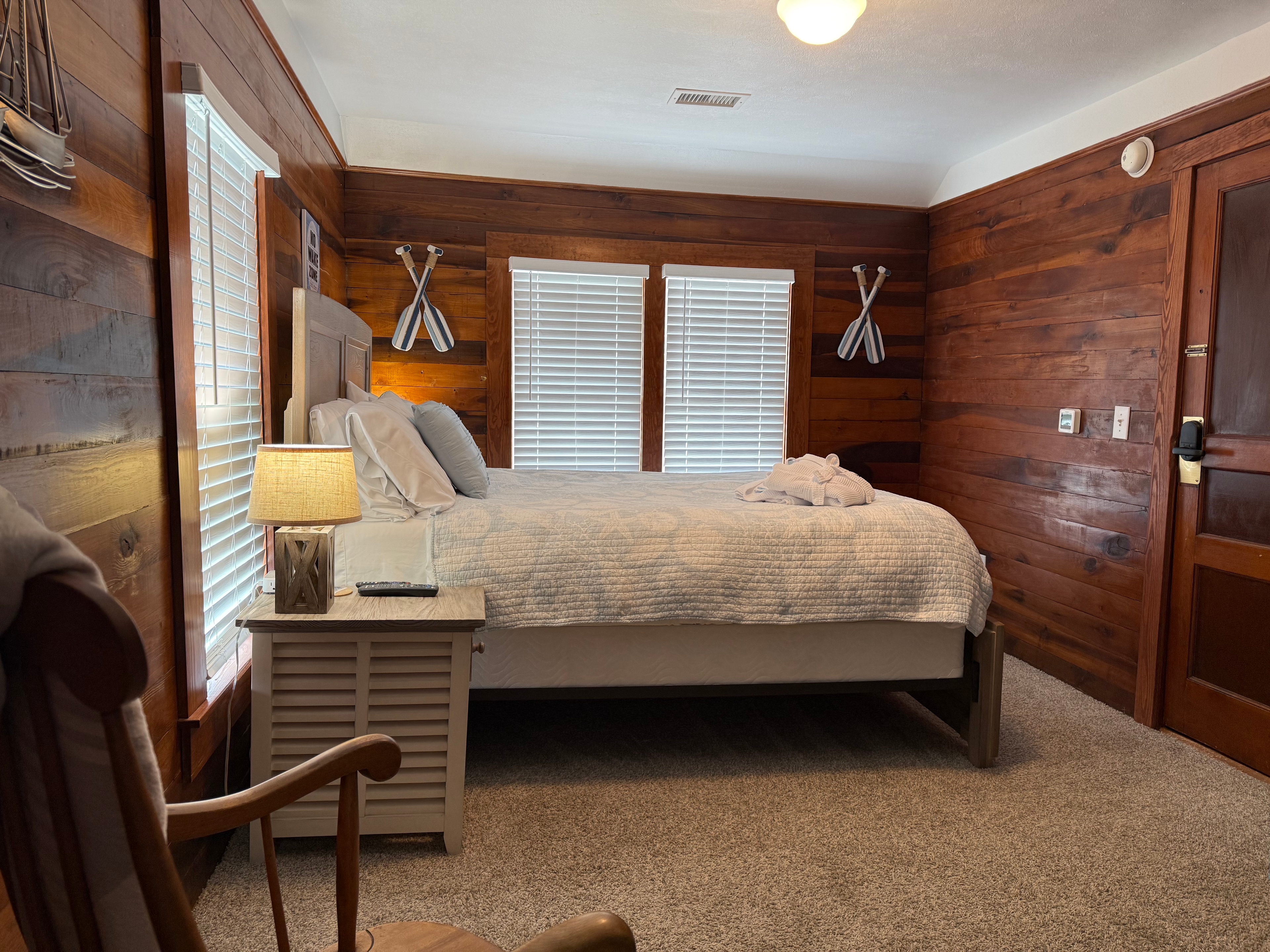 A wide shot of a rustic guest room featuring warm, natural wood-paneled walls and a bed with a white comforter. The room is decorated with nautical touches, including silver oars mounted on the walls, louvered bedside tables with lamps, and a wooden rocking chair in the foreground.