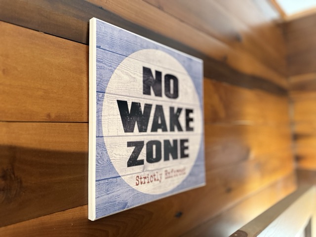 A close-up, angled shot of a square wooden sign mounted on a natural wood-paneled wall. The sign features a circular white background with the bold black text "NO WAKE ZONE" over a light blue wood-grain pattern.