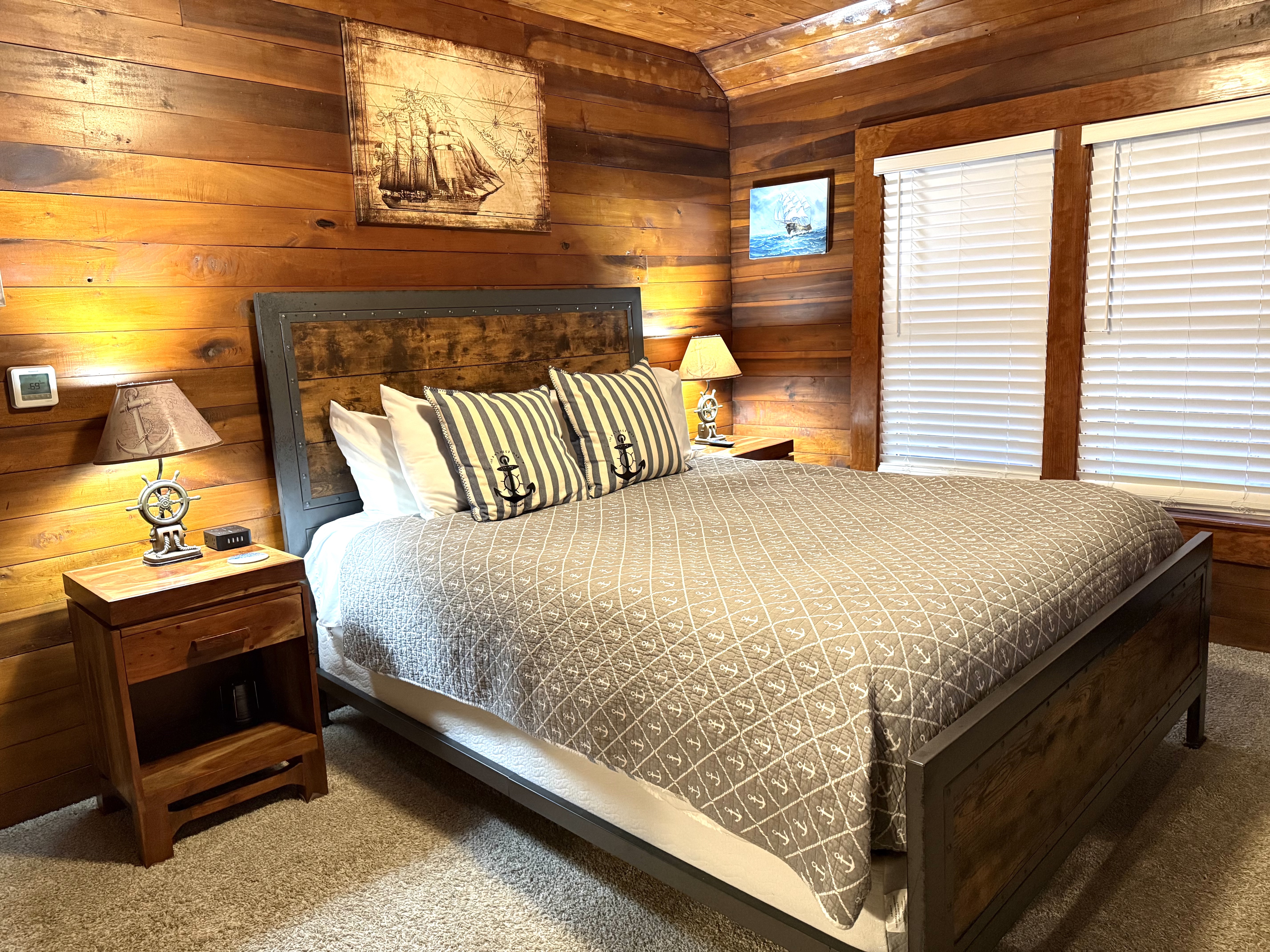 A rustic bedroom featuring horizontal natural wood-paneled walls and a queen bed with a dark wood frame and a gray patterned coverlet. The bed is accented with striped pillows featuring anchor motifs and is flanked by matching wood nightstands with nautical-themed lamps. A large vintage-style illustration of a sailing ship hangs on the wall above the headboard.