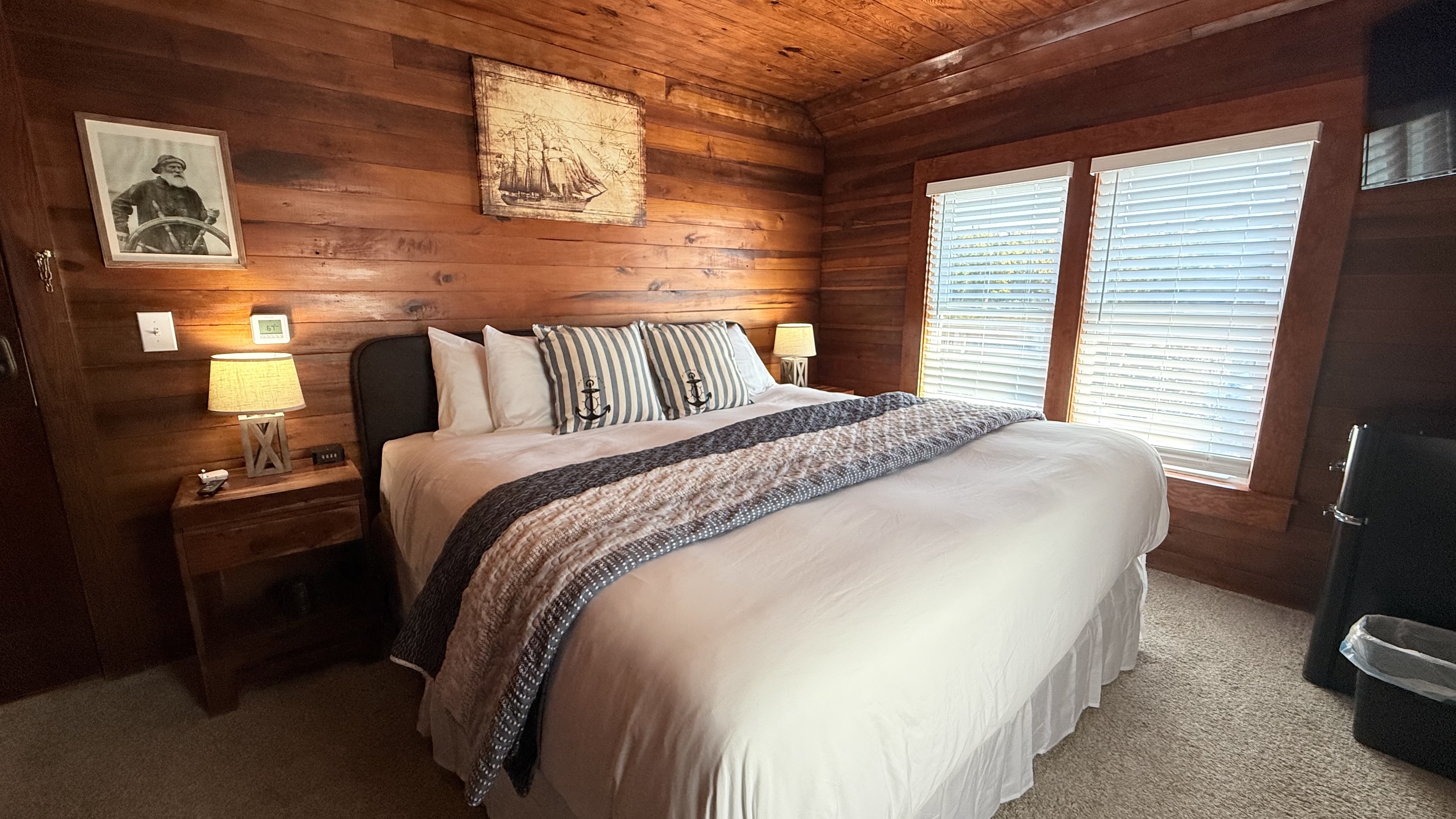 A rustic bedroom featuring horizontal natural wood-paneled walls and a queen bed with a dark wood frame and a gray patterned coverlet. The bed is accented with striped pillows featuring anchor motifs and is flanked by matching wood nightstands with nautical-themed lamps. A large vintage-style illustration of a sailing ship hangs on the wall above the headboard.
