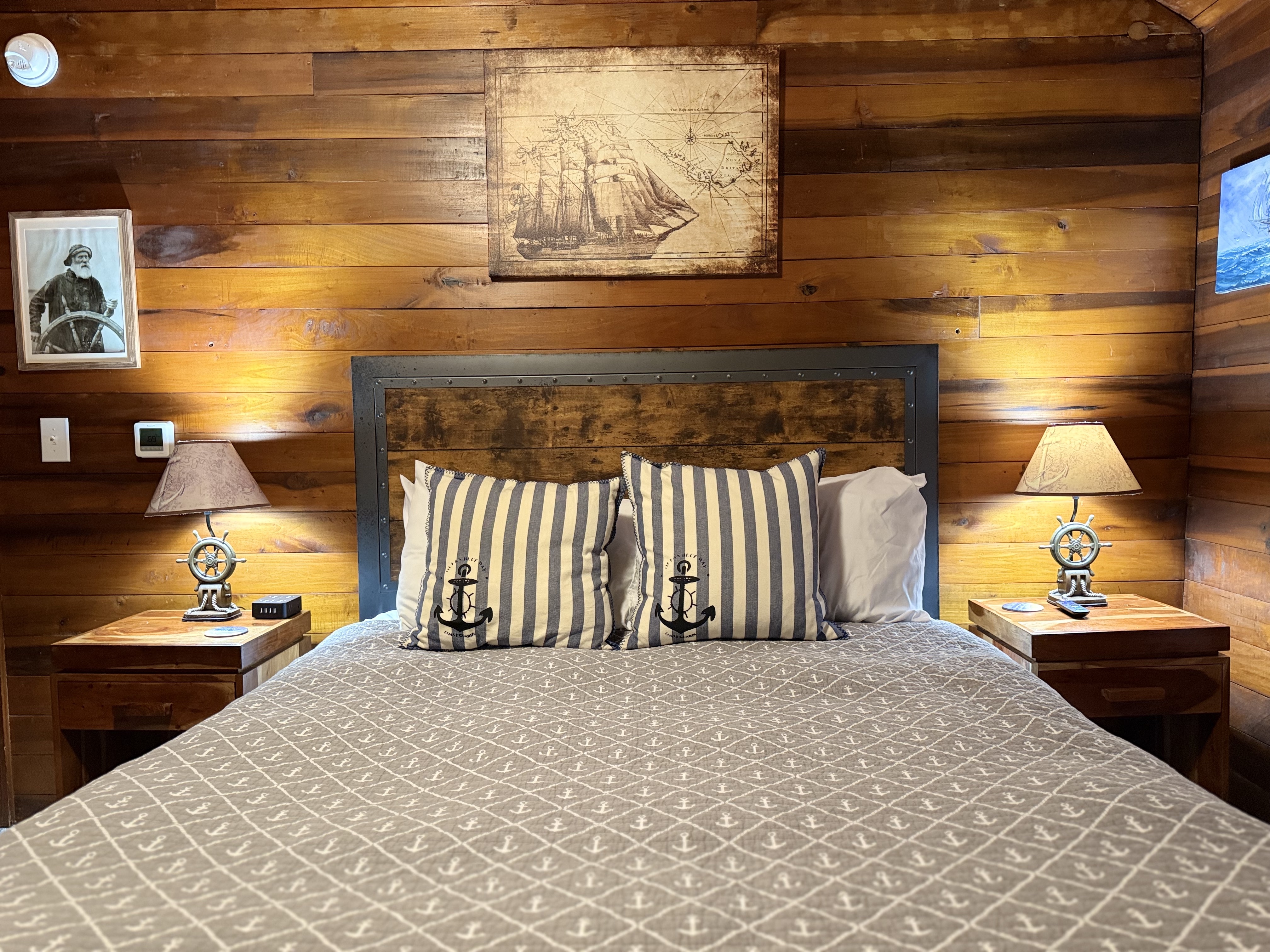 A guest bed featuring a large wooden headboard and a gray patterned coverlet, accented with striped pillows displaying anchor motifs. The room has rich natural wood-paneled walls and is decorated with vintage nautical art, including a large illustration of a sailing ship and a portrait of a sailor.