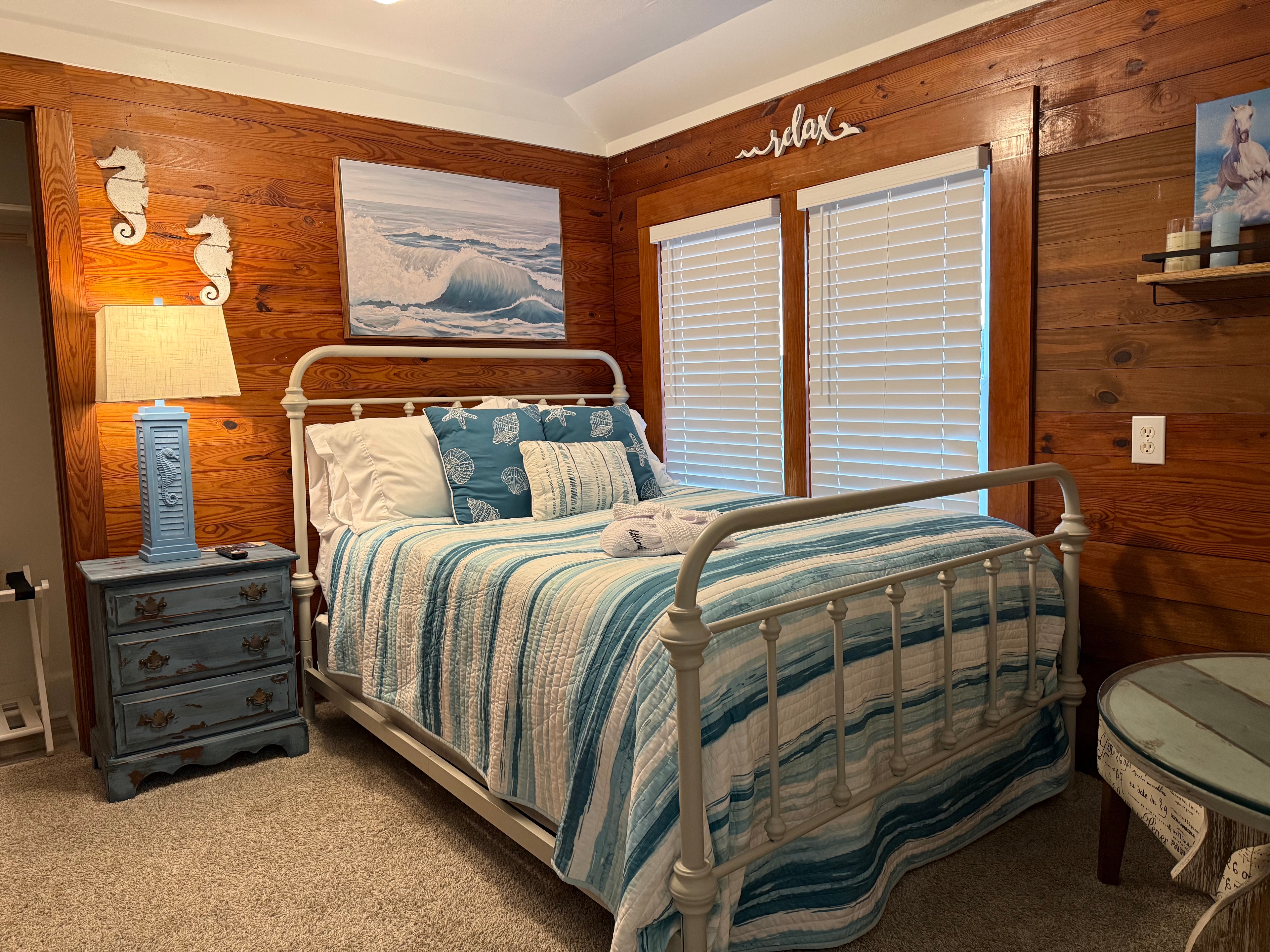 A cozy guest room featuring a white metal bed frame with blue and white striped bedding. The room has warm, natural wood-paneled walls decorated with coastal-themed art, including two white seahorses and a large painting of a breaking ocean wave. A light blue bedside lamp stands on a matching blue dresser next to the bed.