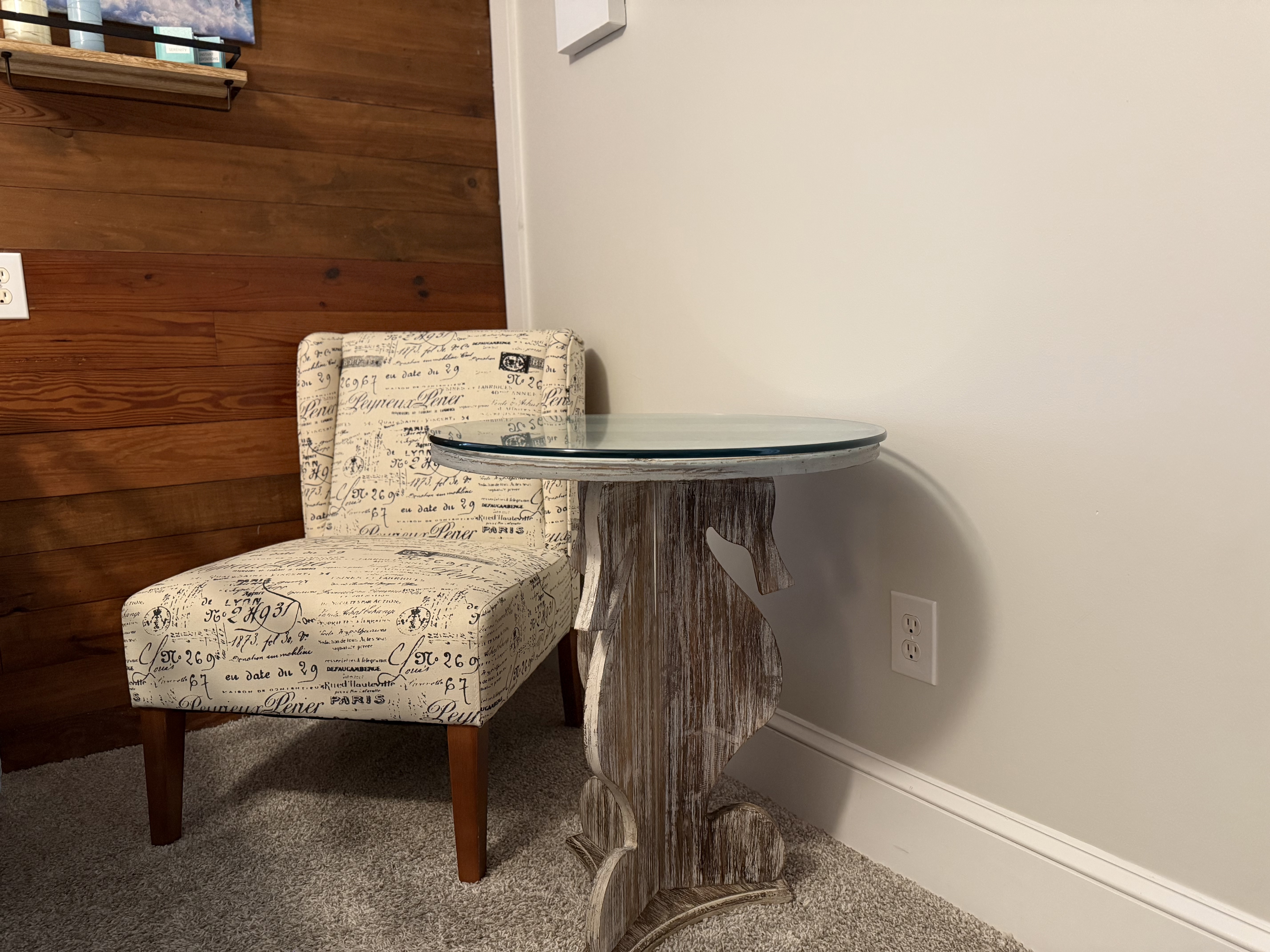 A cozy seating area in a guest room featuring a small armchair with vintage-style script upholstery and dark wood legs. Next to the chair is a round glass-top table with a driftwood-inspired base, set against a light gray wall and a natural wood-paneled accent wall.