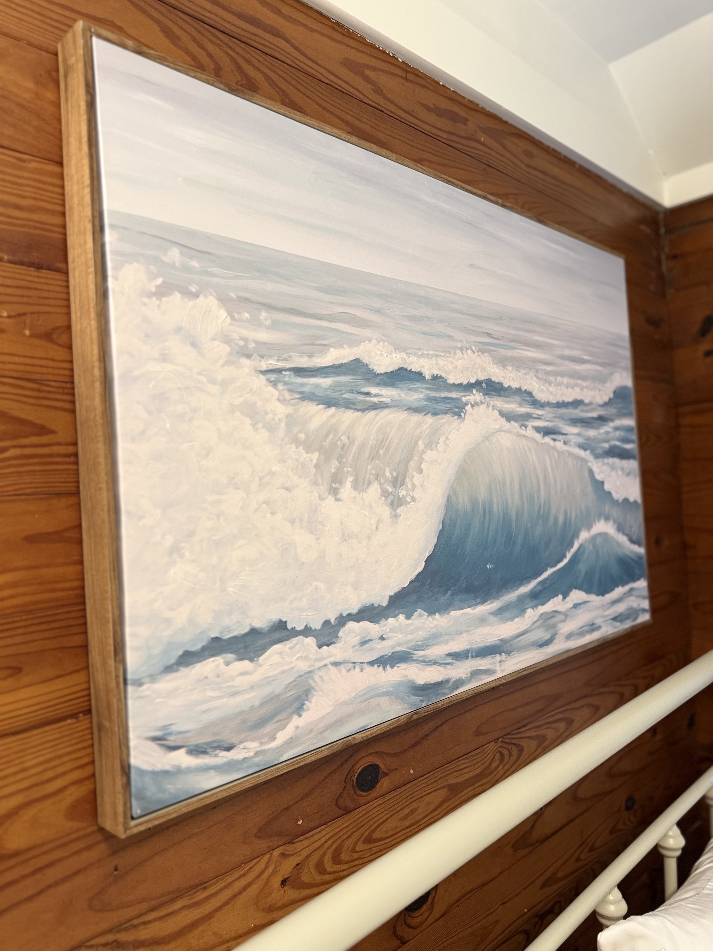 A close-up, angled shot of a large rectangular painting of a cresting ocean wave in shades of blue and white, encased in a thin light-wood frame and mounted on a rustic wood-paneled wall.