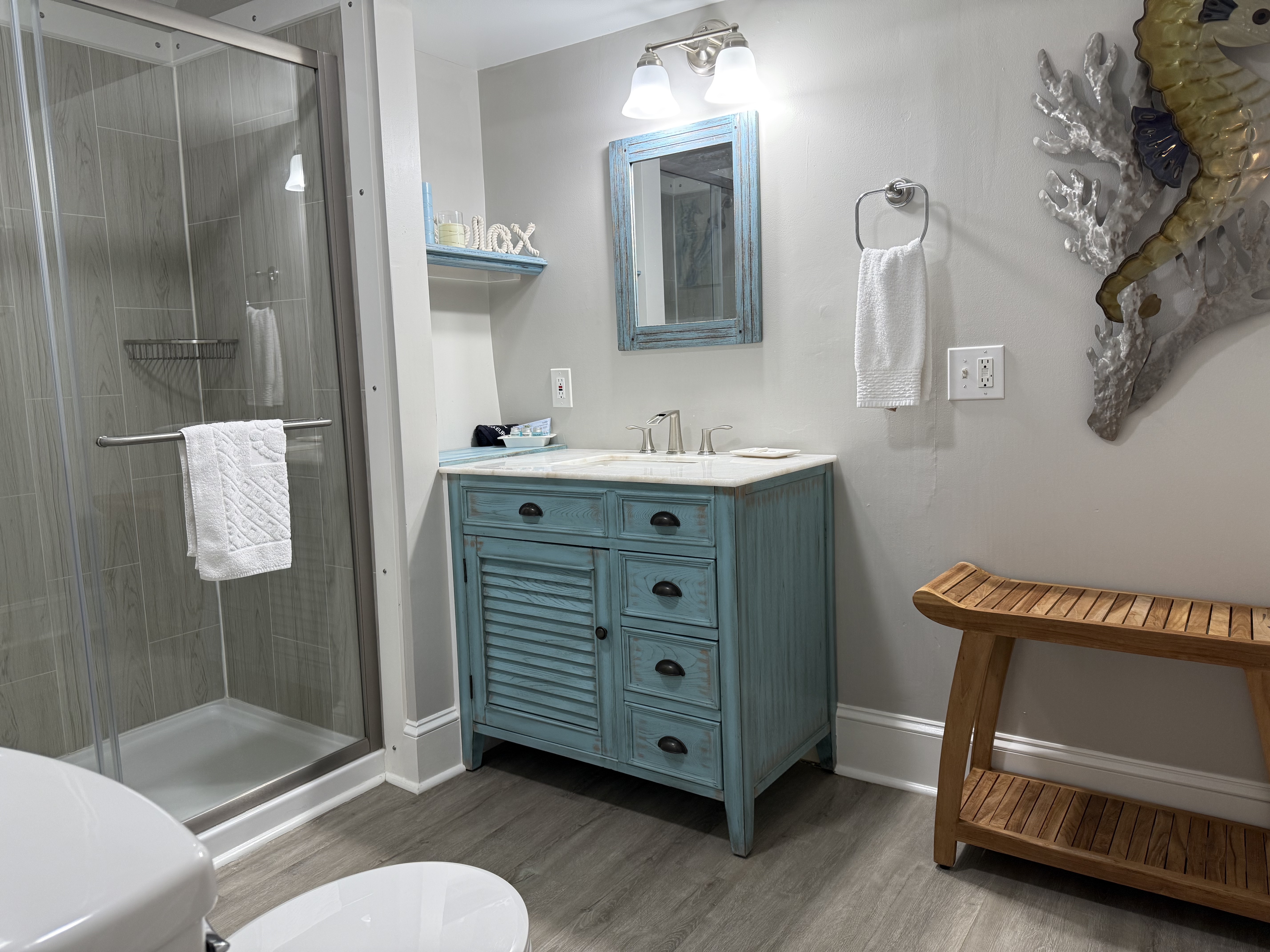 A modern bathroom featuring a light blue louvered vanity with a white countertop and a matching blue-framed mirror. The room includes a glass-enclosed walk-in shower with wood-style tile, a small wooden bench, and light gray walls decorated with a large three-dimensional seahorse sculpture.