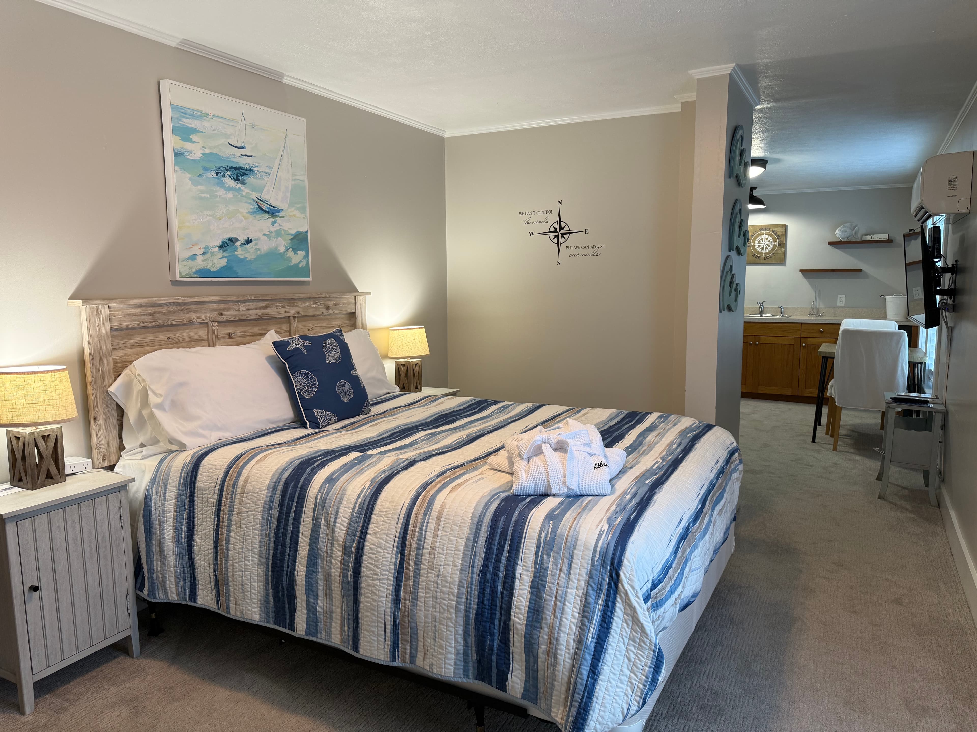 A spacious bedroom at The Atlantic Inn featuring a bed with blue and white striped bedding, white bathrobes, and coastal decor, with a view into an adjoining kitchenette and living area.
