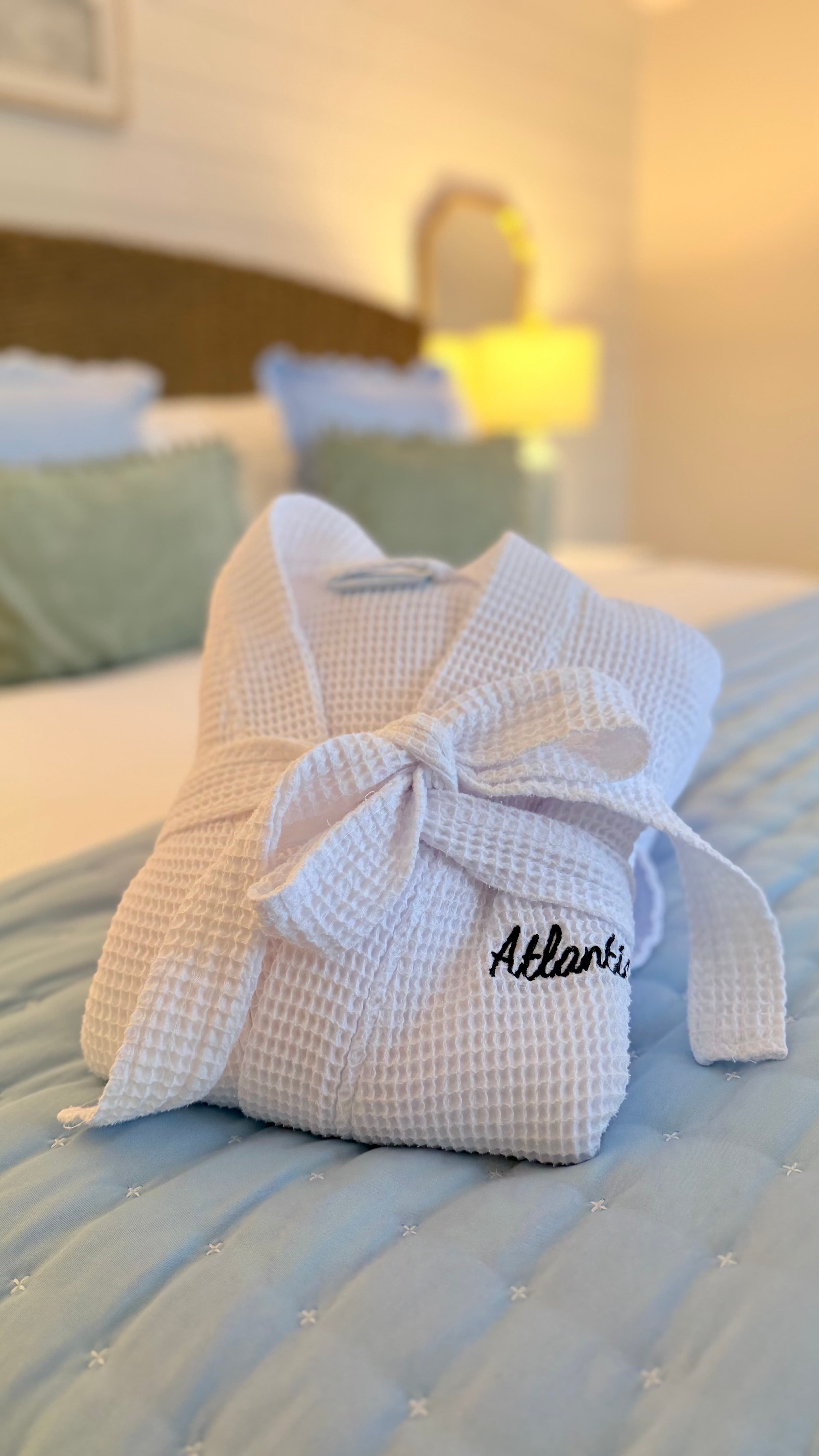 A close-up of two white waffle-weave bathrobes embroidered with "Atlantic" in black script, neatly folded on a bed with blue and white striped bedding.