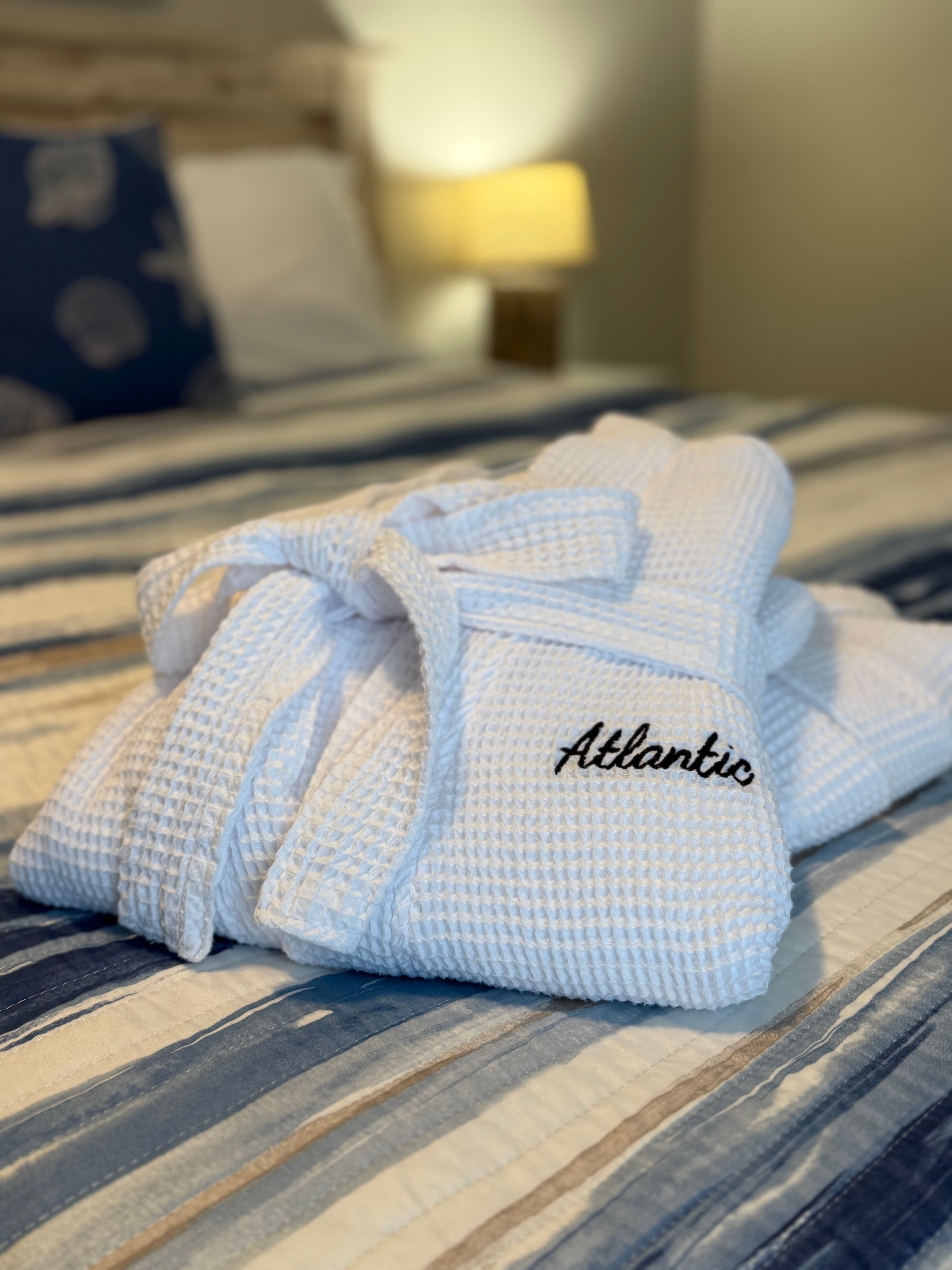 A close-up of two white waffle-weave bathrobes embroidered with "Atlantic" in black script, neatly folded on a bed with blue and white striped bedding.