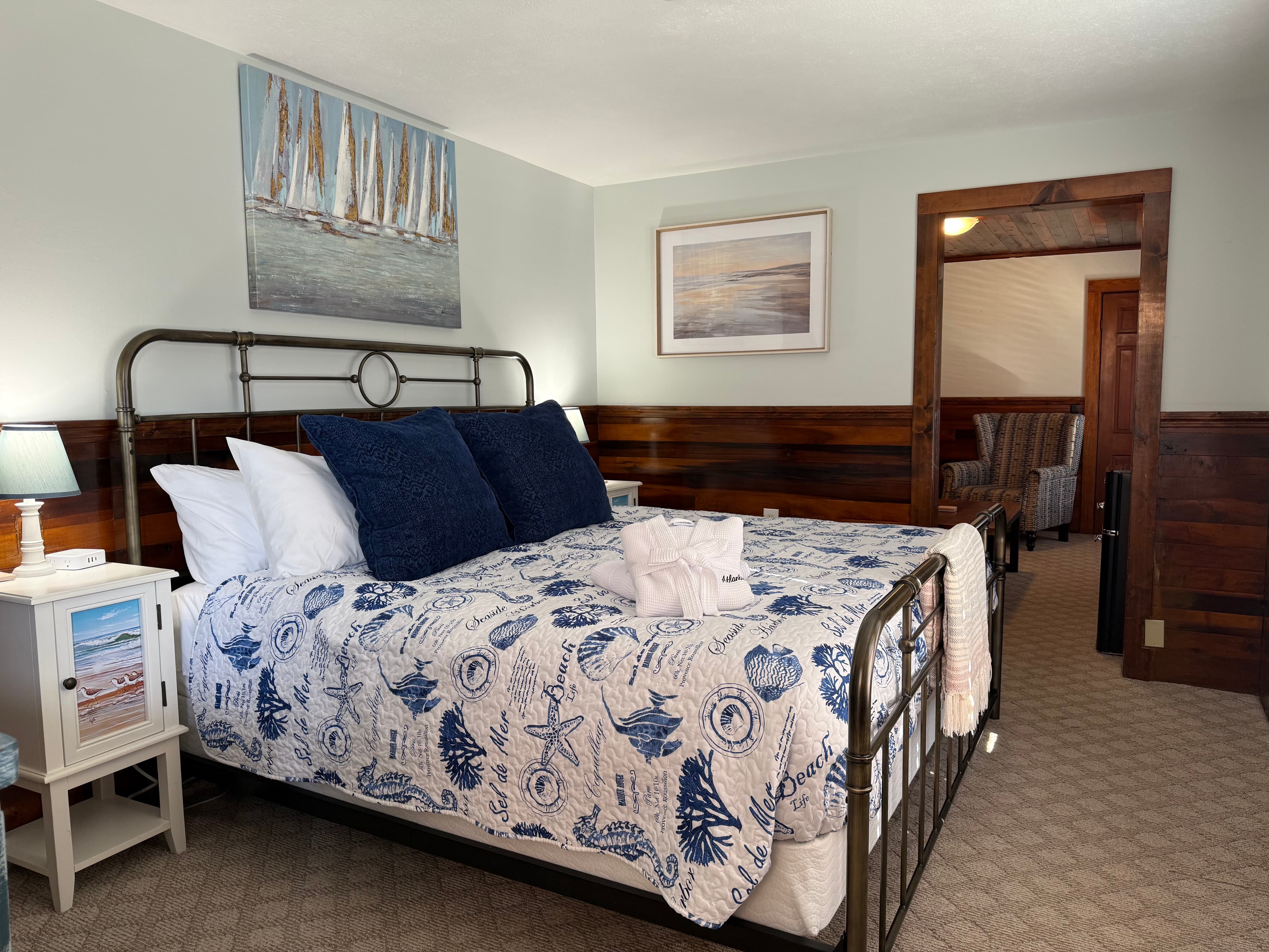 A rustic bedroom at The Atlantic Inn featuring a large bed with nautical seashell-patterned bedding and an iron headboard, set against walls with original dark wood paneling and coastal artwork.