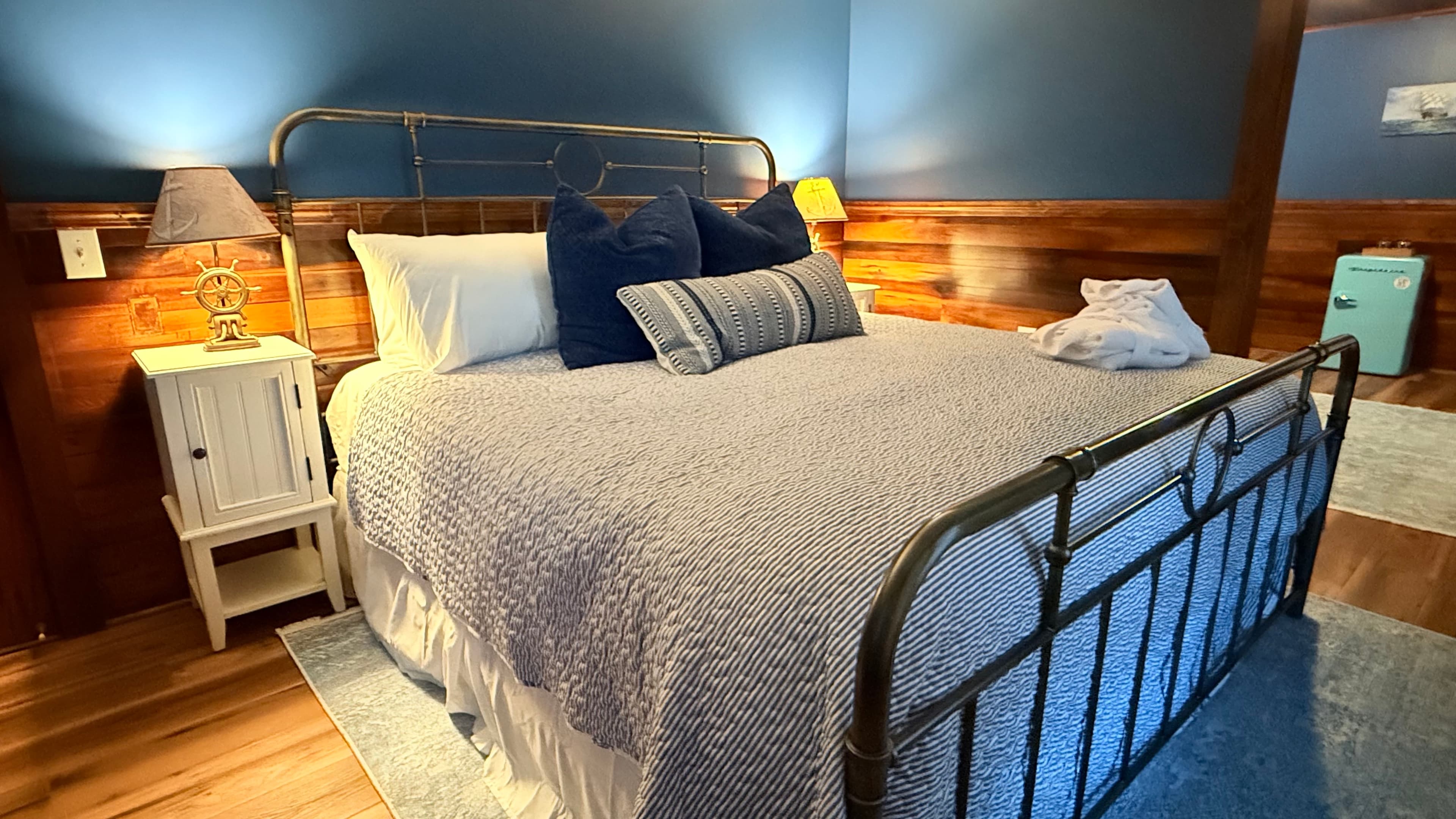 A rustic bedroom at The Atlantic Inn featuring a large bed with nautical seashell-patterned bedding and an iron headboard, set against walls with original dark wood paneling and coastal artwork.