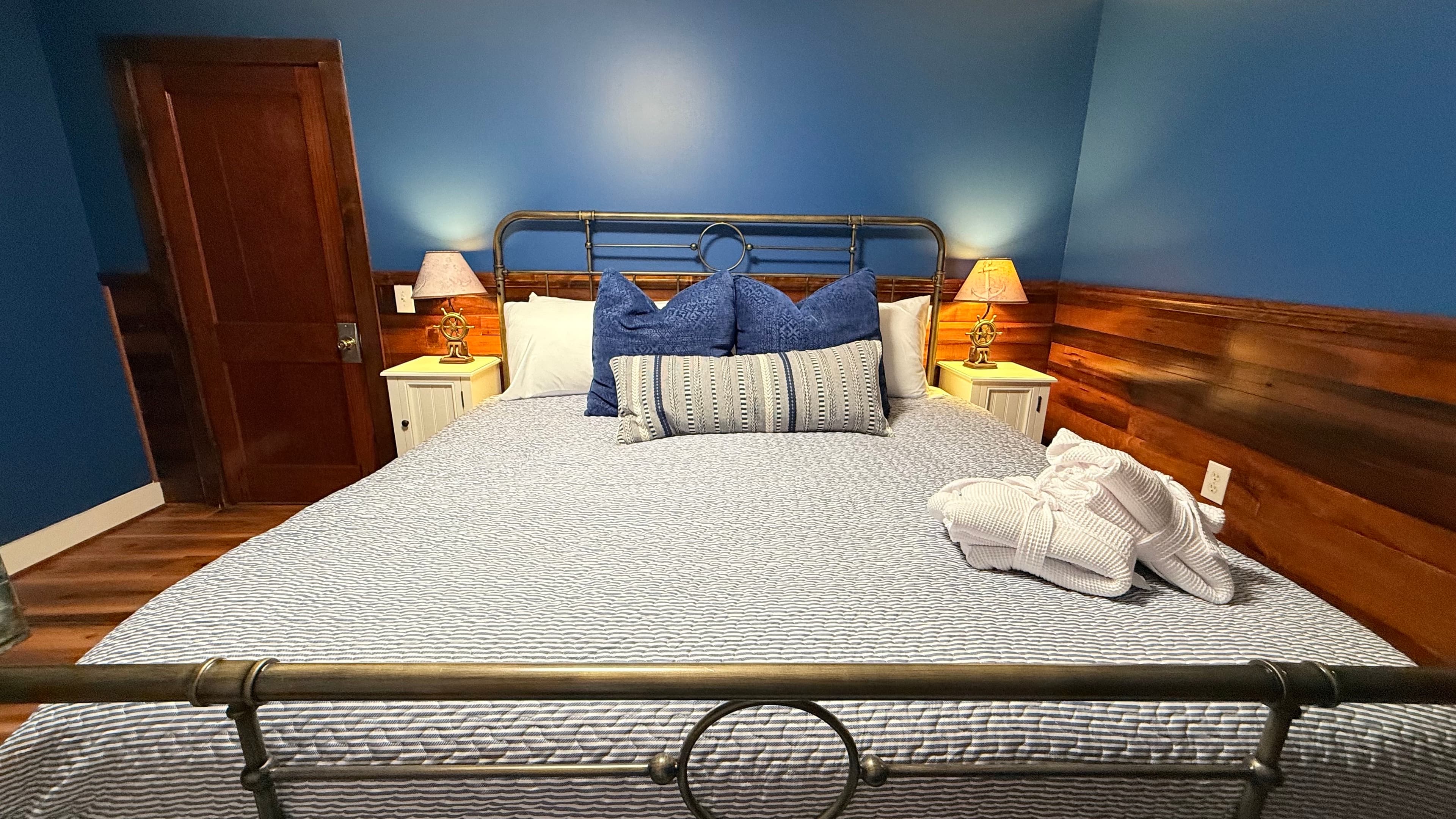 A nautical-themed bedroom at The Atlantic Inn featuring a large bed with blue and white seashell-patterned bedding and an iron headboard. The room is accented with original dark wood wainscoting, coastal artwork, and white bedside tables with lamps.