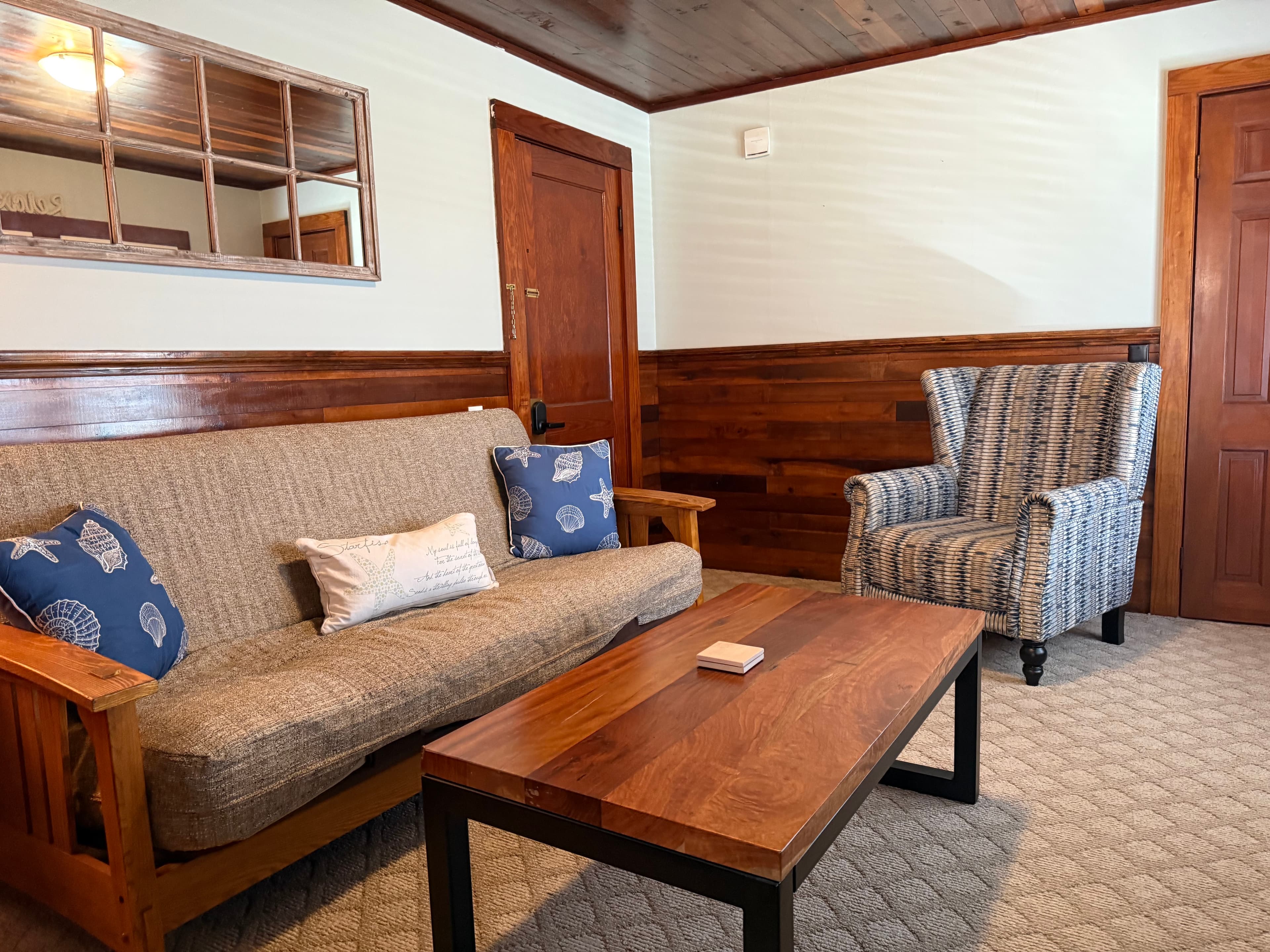 A rustic living area featuring wood wainscoting and a matching dark wood coffee table. The room includes a tan futon with nautical pillows, a striped wingback chair, and classic wooden doors.
