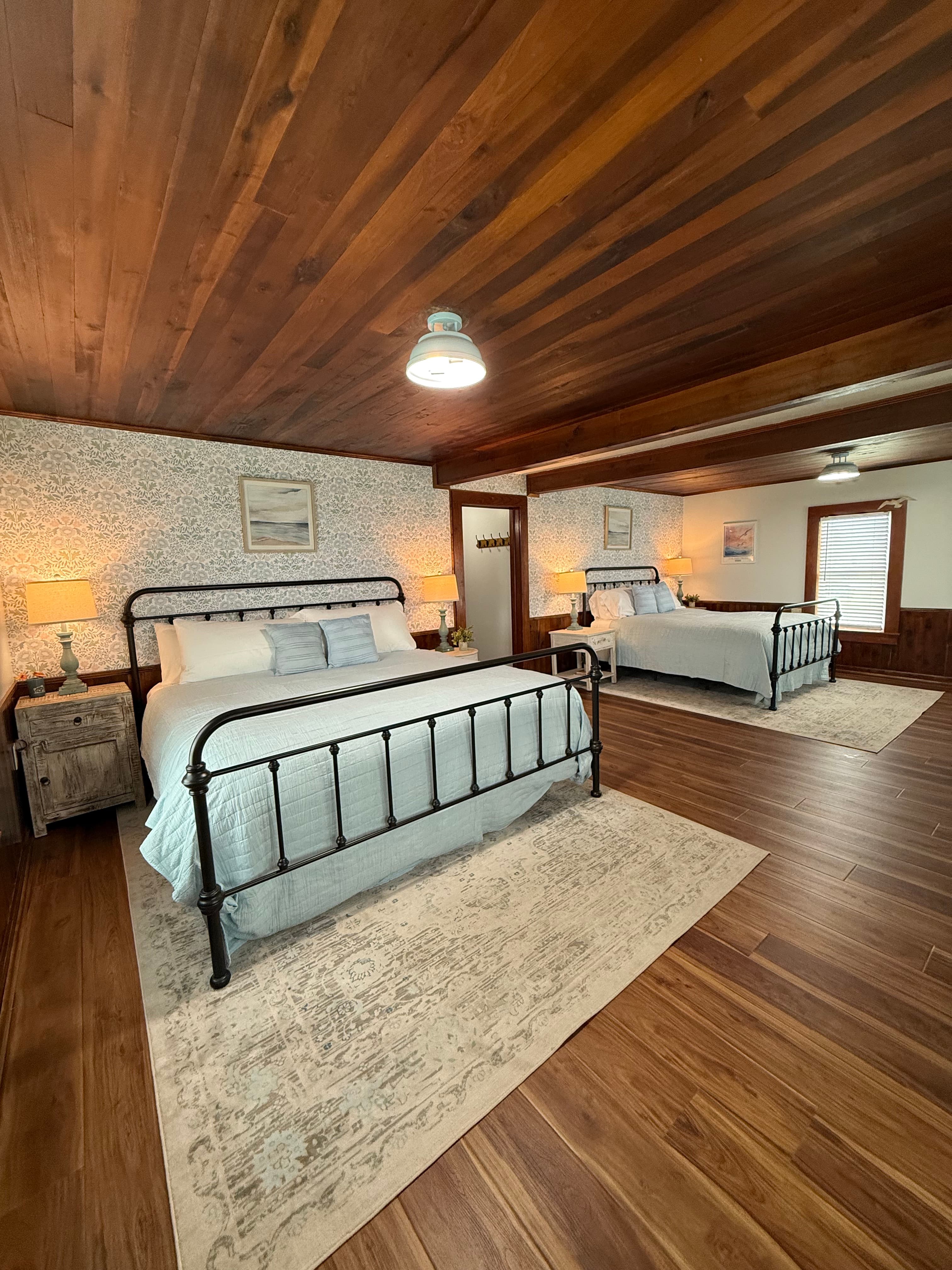 A large, newly remodeled suite featuring a king and a queen bed with iron frames and light blue bedding. The room showcases polished wood floors and a stunning original wood ceiling, accented by coastal wallpaper and warm bedside lighting.