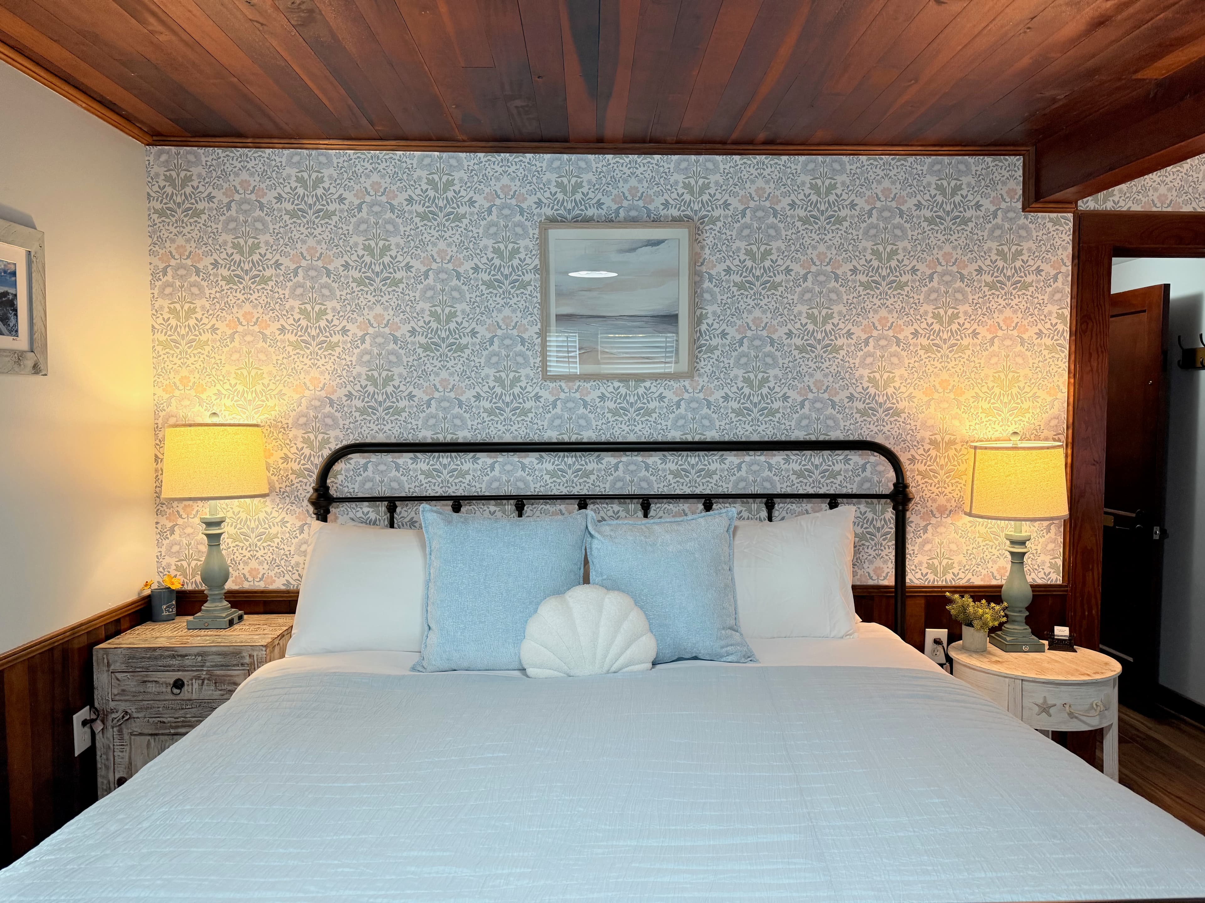 A large, newly remodeled suite featuring a king bed and a queen bed with iron frames and light blue bedding. The room showcases polished wood floors and a stunning original wood ceiling, accented by coastal wallpaper and warm bedside lighting.