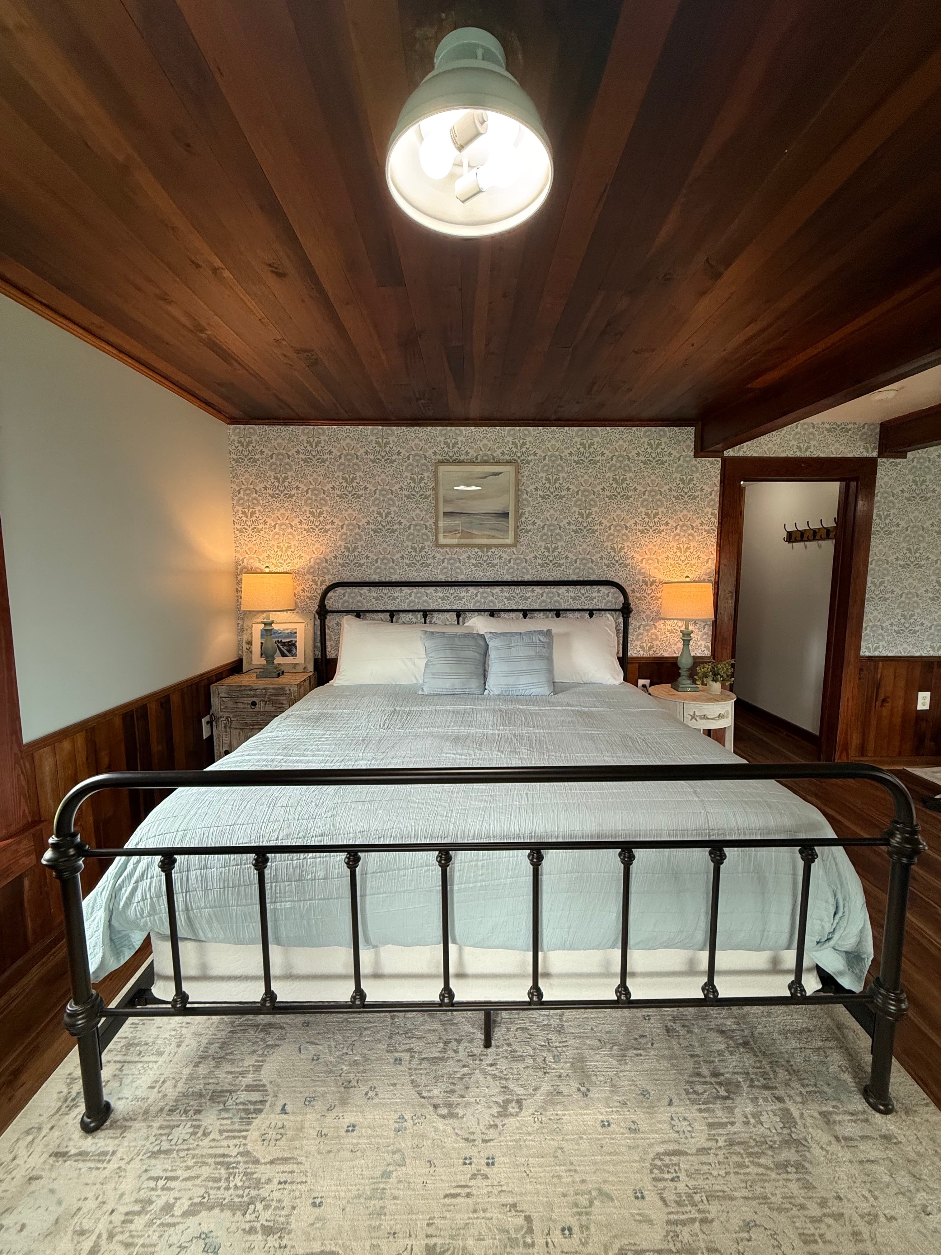 A large, newly remodeled suite featuring a king bed and a queen bed with iron frames and light blue bedding. The room showcases polished wood floors and a stunning original wood ceiling, accented by coastal wallpaper and warm bedside lighting.