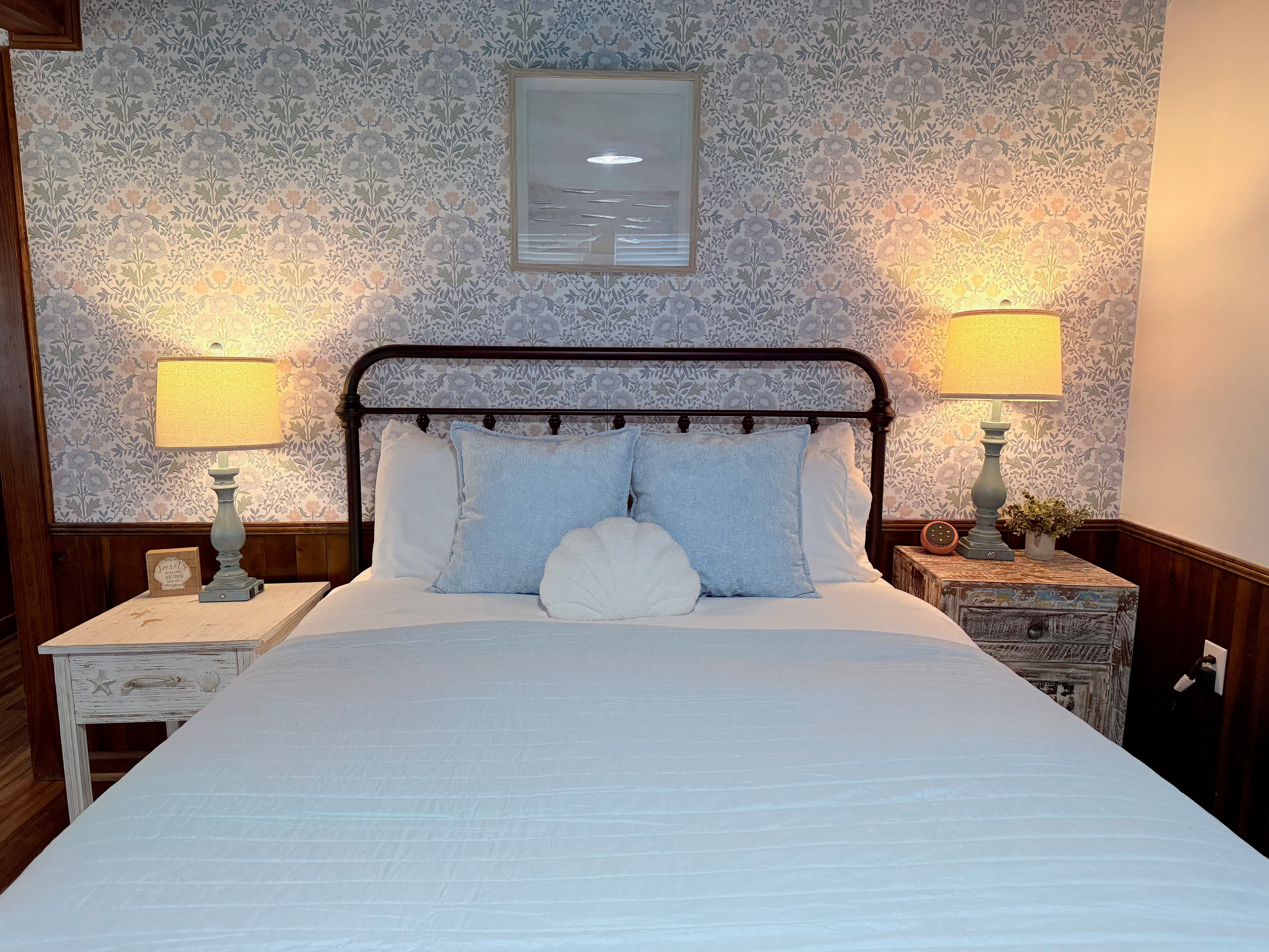 A detail view of a bedroom in a newly remodeled suite featuring a queen-size bed with an iron frame and light blue bedding. The room is decorated with coastal-patterned wallpaper, a rustic wooden nightstand with a lamp, and original dark wood ceilings and flooring.